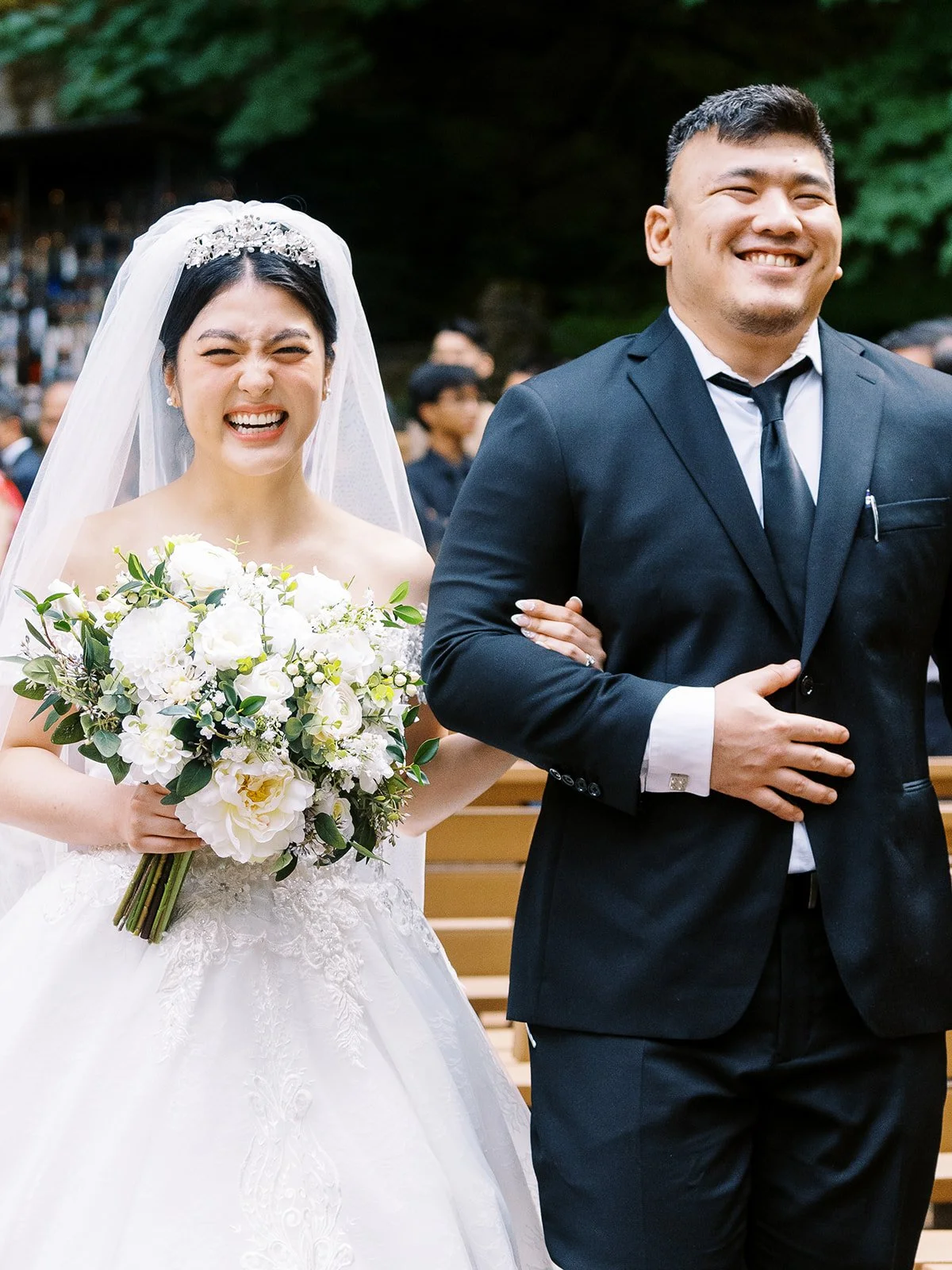 Close-up of the bride laughing joyfully beside a smiling man in a black suit as they walk back up the aisle at The Grotto, her white peony bouquet in hand
