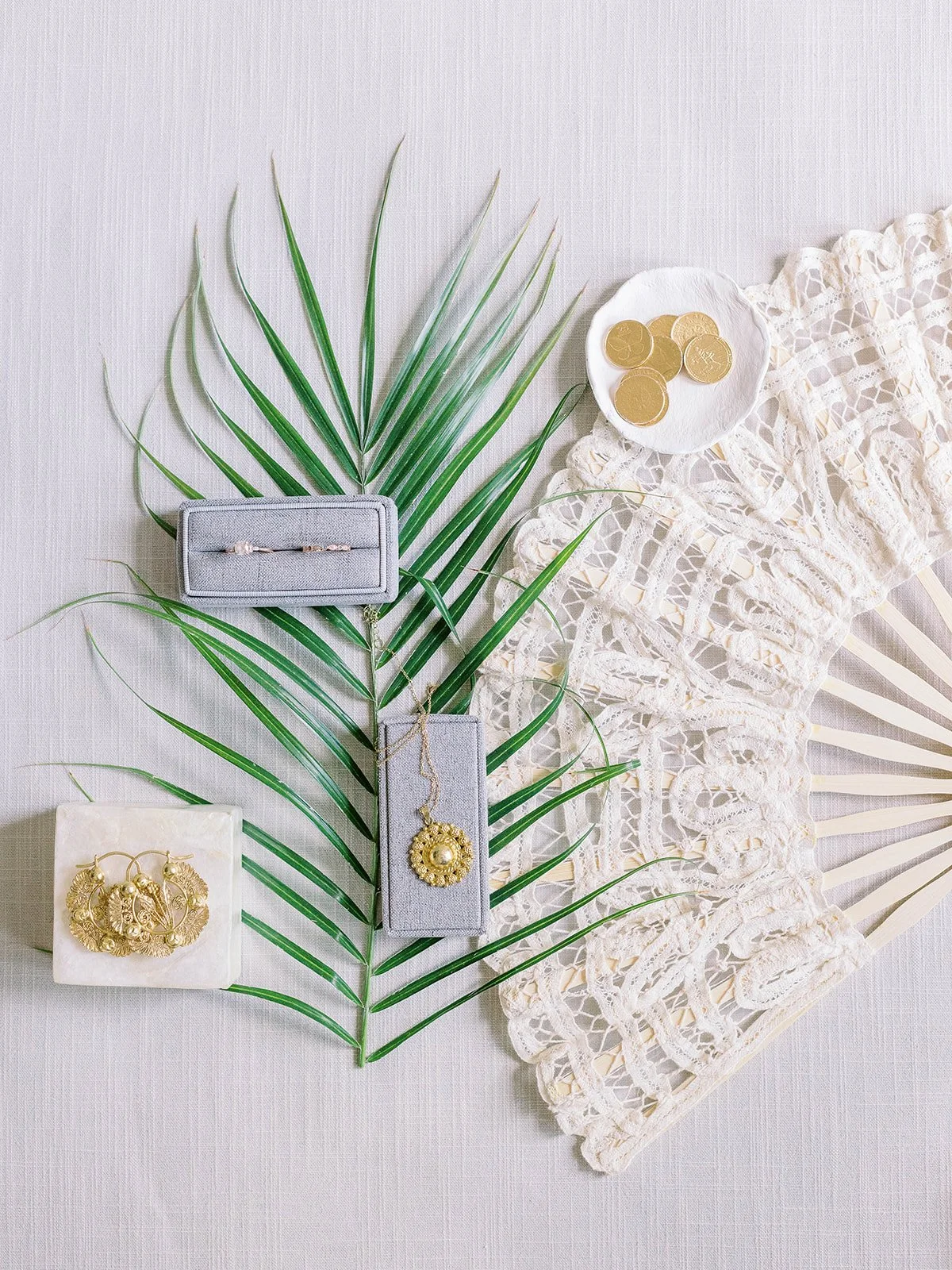 Wedding flat lay with gold jewelry, a ring box, lace hand fan, tropical palm leaf, and gold coins on a white linen surface