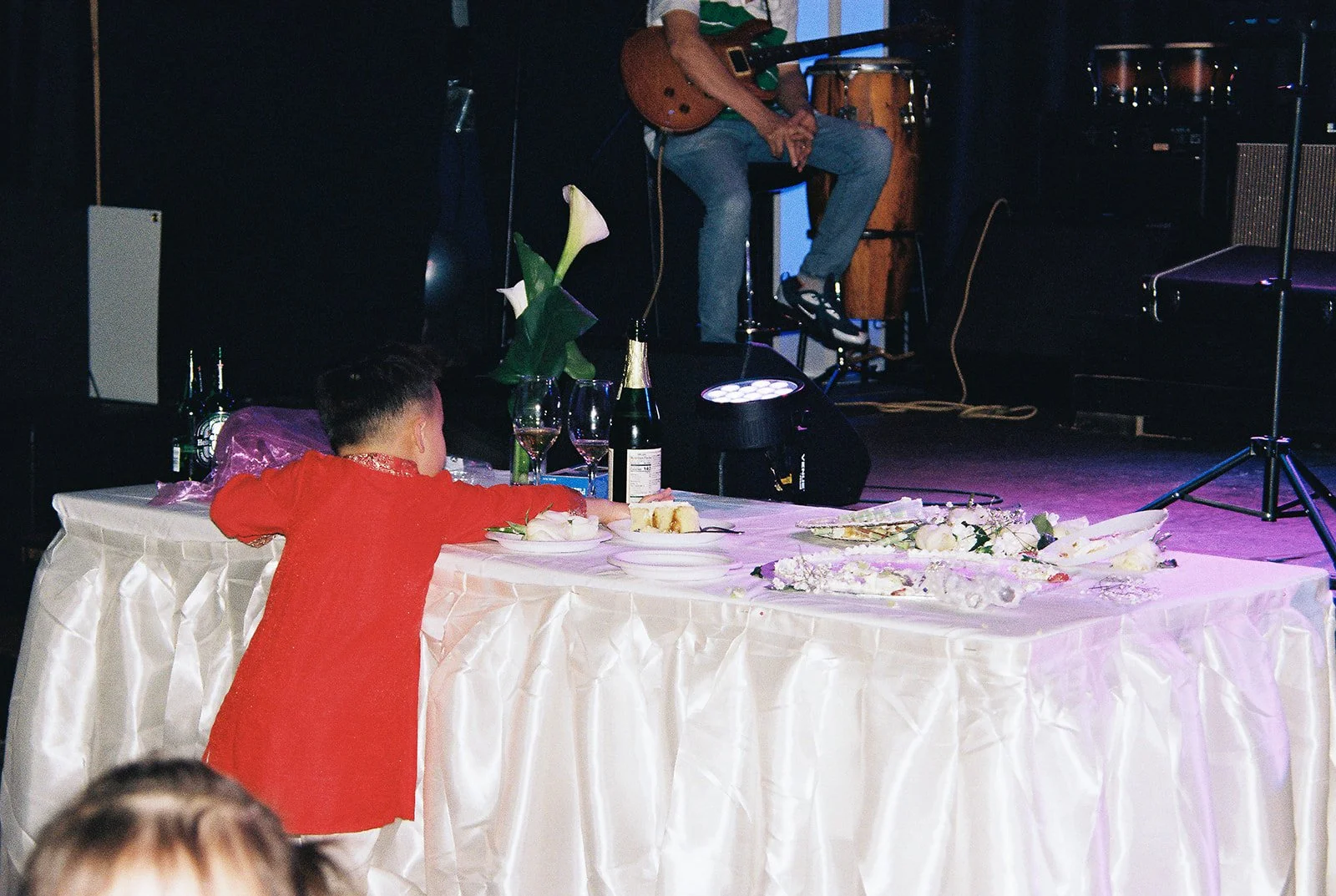Candid of a child in his red áo dài leaning on the sweetheart table at the Mekong Bistro reception, a guitarist visible on stage above him