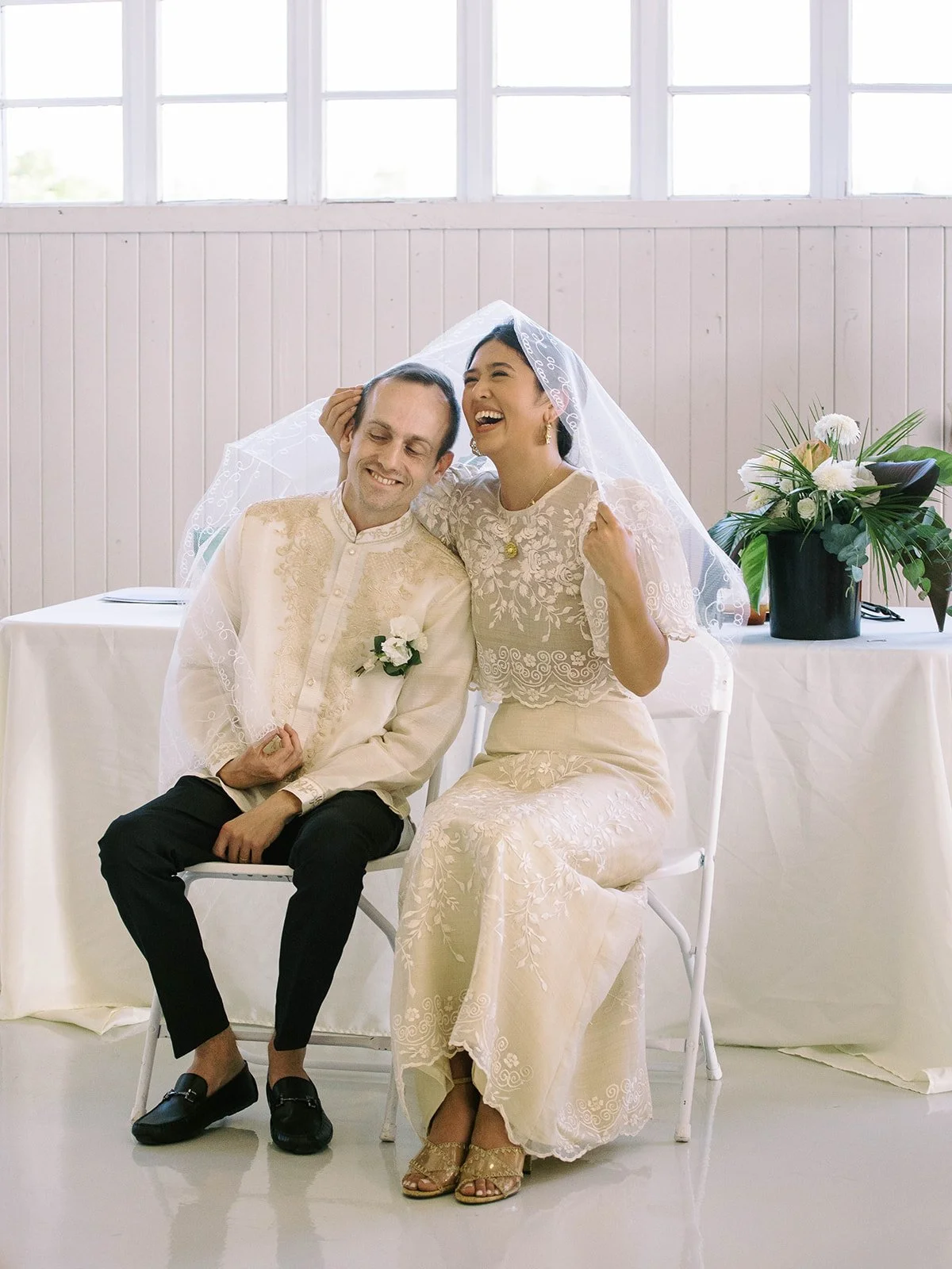 Bride and groom laughing together under the traditional Filipino wedding veil during their indoor ceremony at Pearson Air Museum
