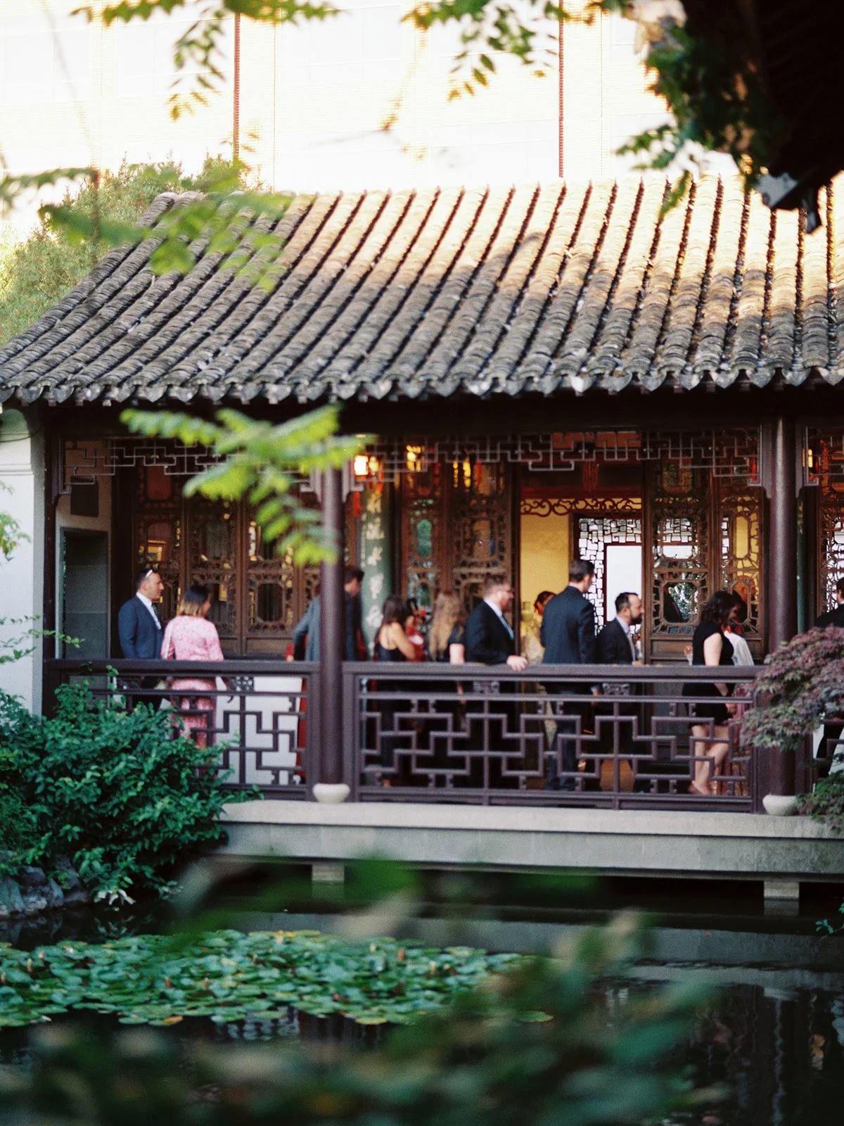 Soft-focus view of guests gathered on the pavilion terrace across the lily pond at golden hour, framed by foreground foliage.