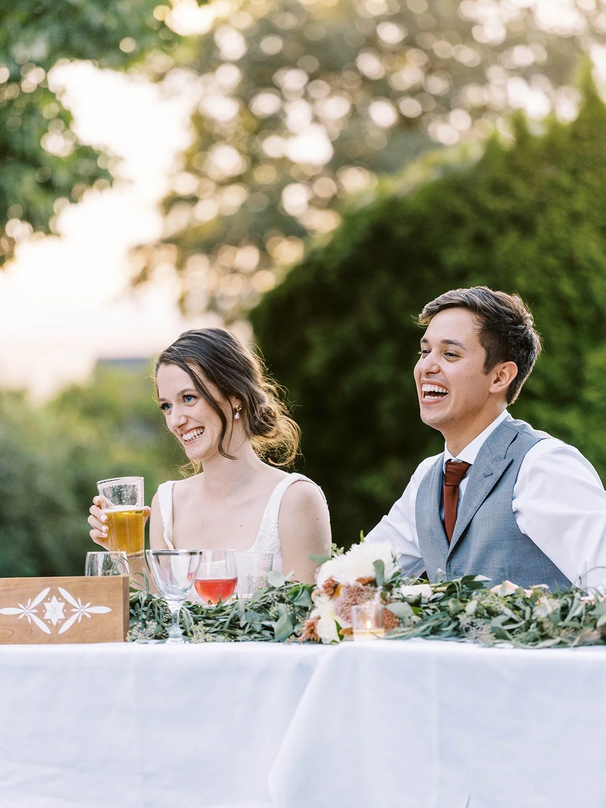 Bride and groom seated at reception table, smiling and holding drinks with greenery and floral decor