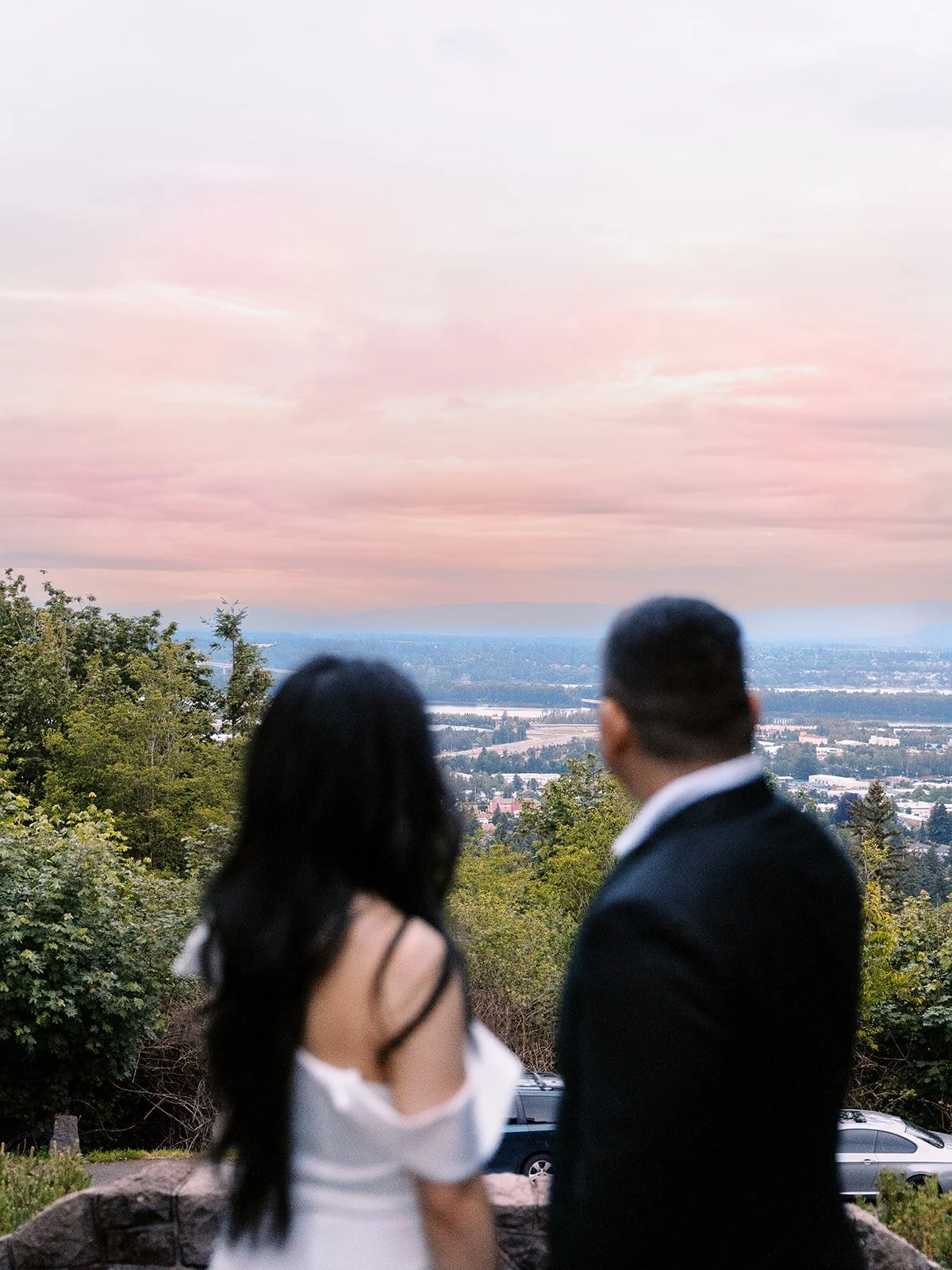 Soft-focus shot of the bride and groom seen from behind, looking out over a pink sunset sky and the Columbia River from The Grotto's upper overlook