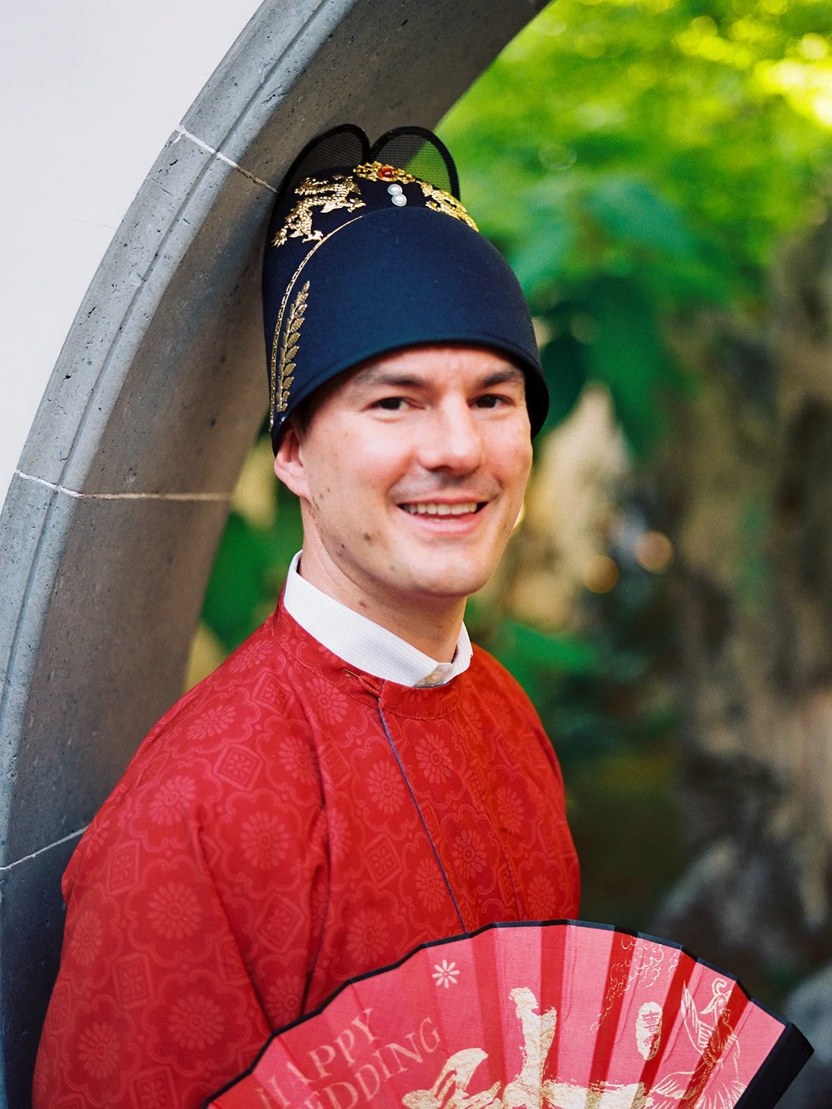 Groom smiling in a red traditional Chinese wedding robe and ornate black ceremonial hat, holding a red "Happy Wedding" fan in a stone moon gate