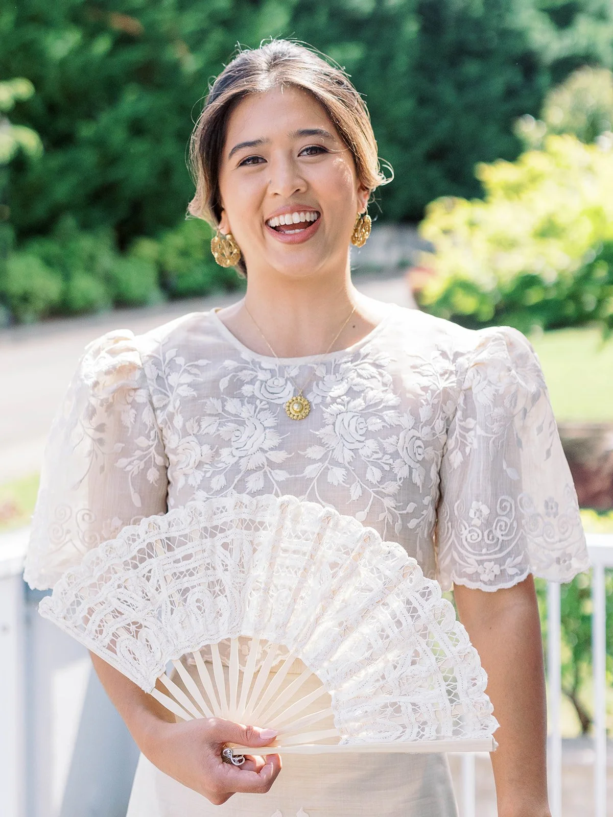 Bride laughing joyfully while holding a white lace fan, wearing a floral embroidered Filipino gown and gold earrings

