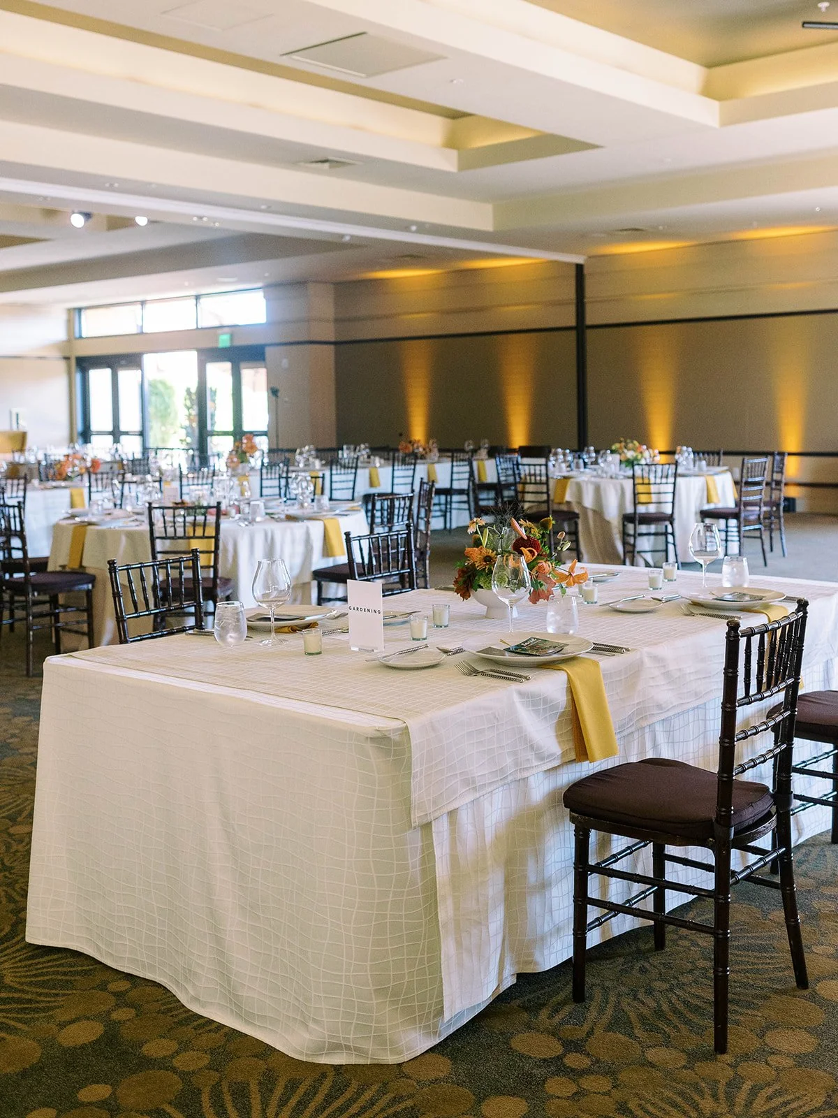Wedding reception ballroom with round and rectangular tables set with white linens, yellow napkins, dark chiavari chairs, and colorful floral centerpieces under warm amber uplighting