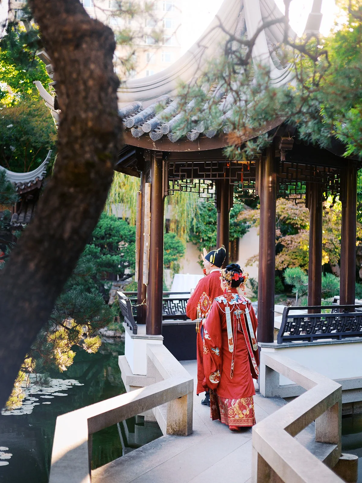 Bride and groom in red traditional Chinese wedding robes walk along a stone pathway beside a lily pond, framed by a pine tree and dark wood hexagonal pavilion at Lan Su Chinese Garden.