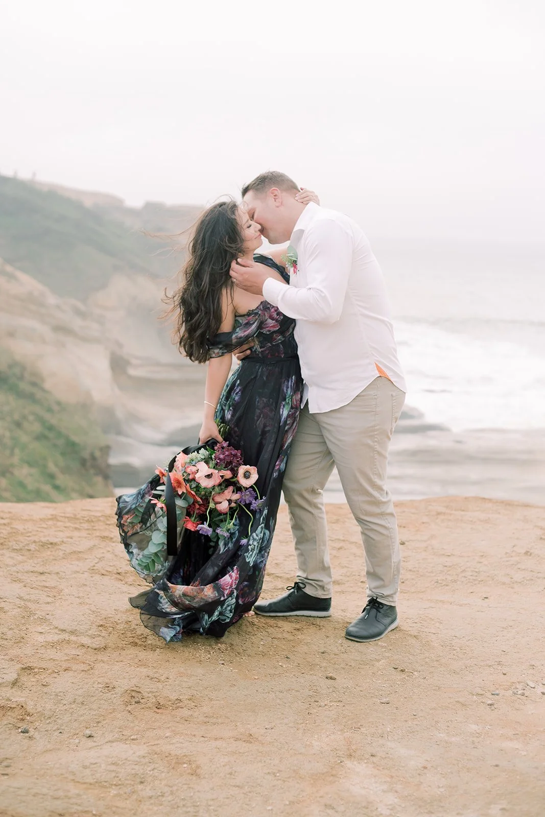 Couple kissing on a cliffside overlooking the Oregon Coast, woman holding a colorful bouquet, soft coastal fog