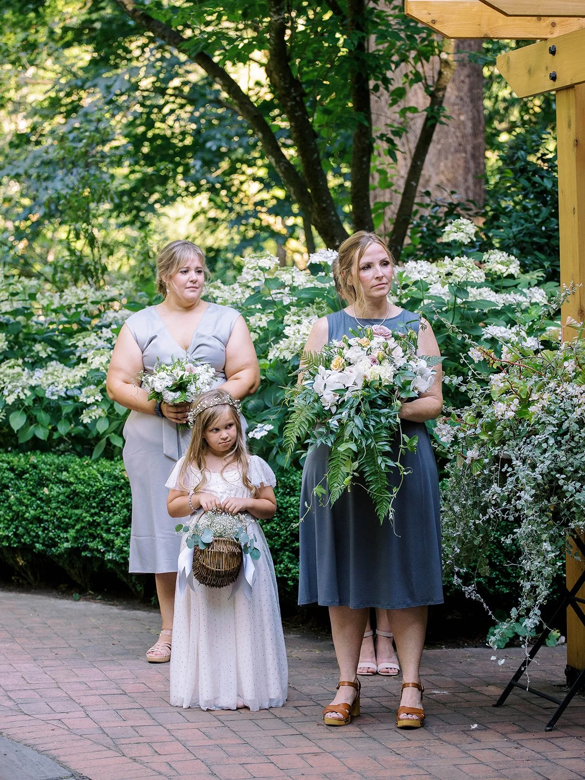 Bridesmaids and a flower girl hold bouquets beneath a wooden pergola surrounded by hydrangeas.