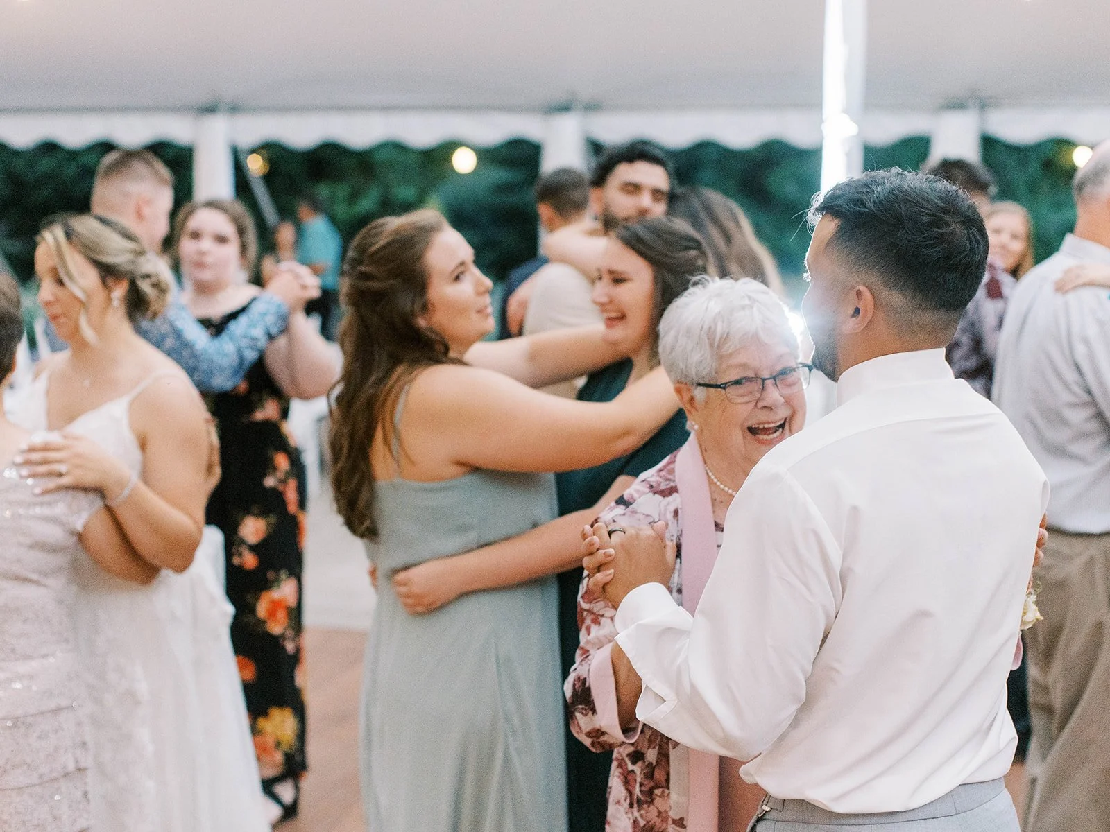 Groom dances with an older guest while friends and family dance nearby at the wedding reception.