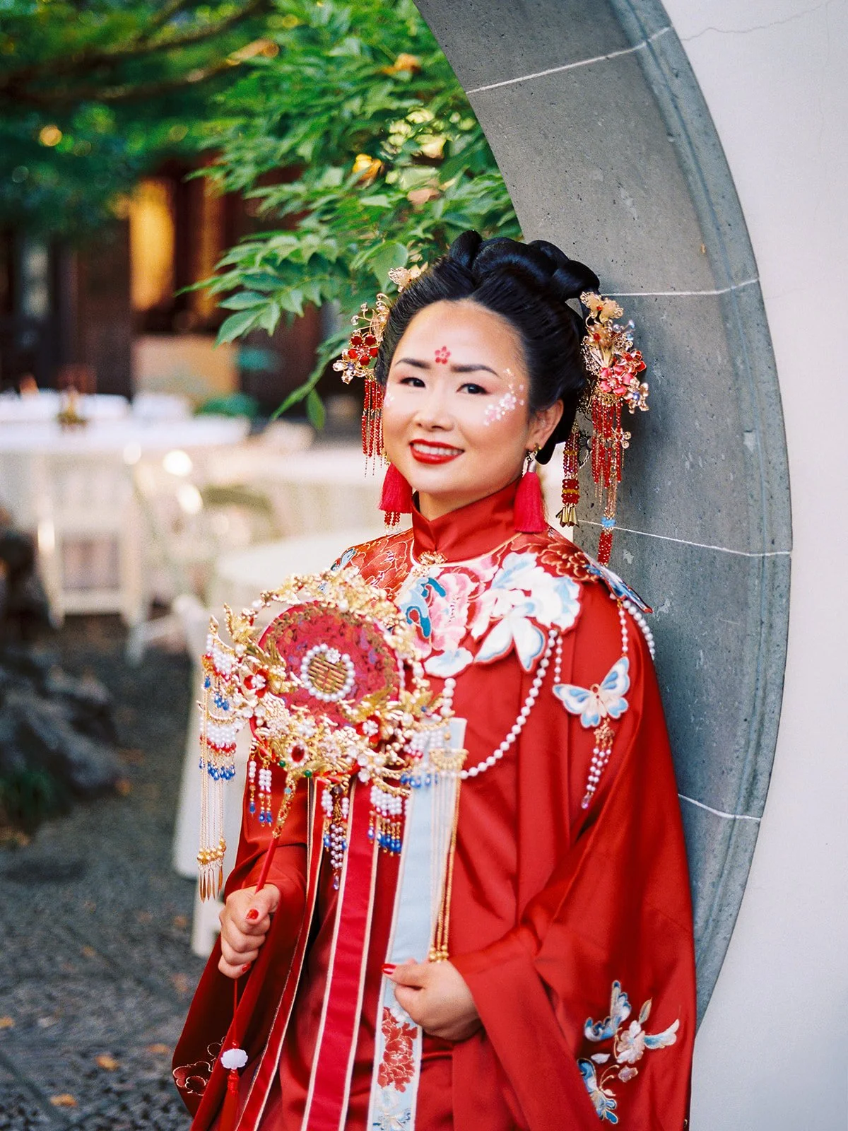 Bride in red embroidered qun kwa wedding gown holding a gold and jeweled bridal bouquet, leaning against a stone moon gate at Lan Su Chinese Garden in Portland