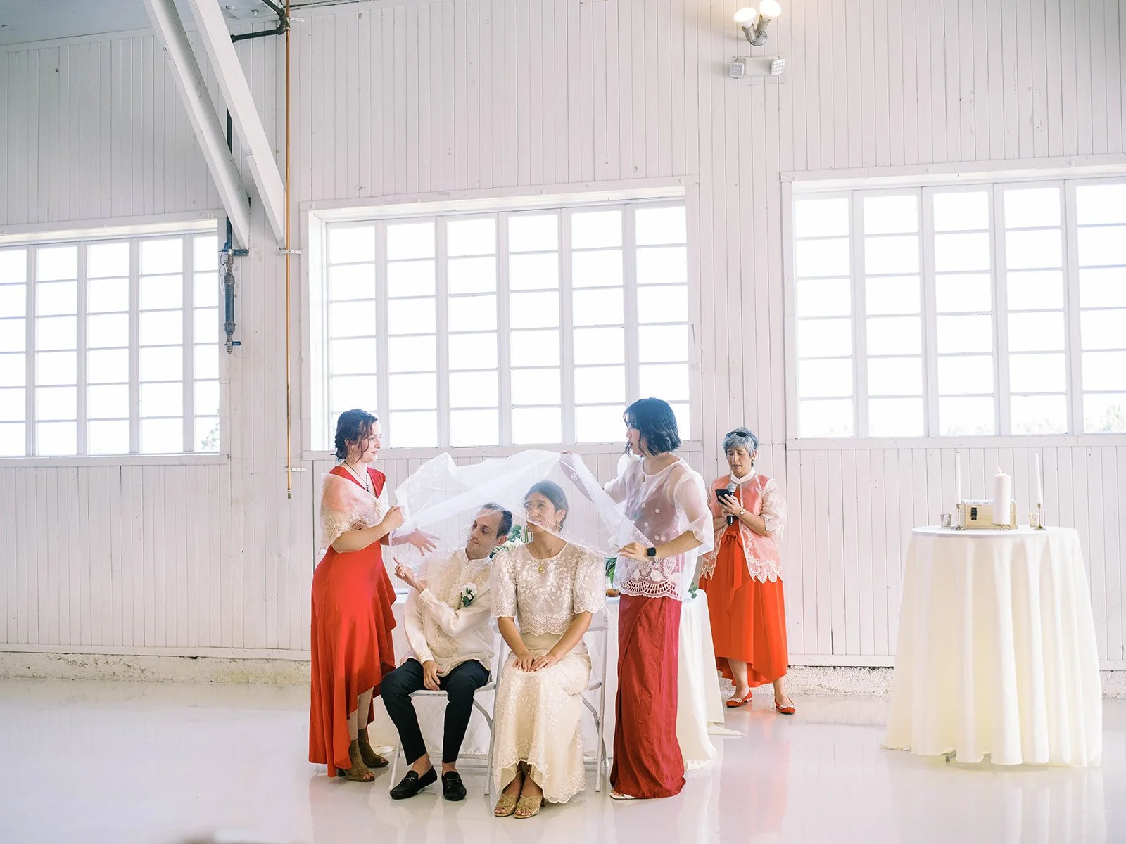 Wedding party members draping the traditional lace veil over the seated bride and groom during the Filipino wedding ceremony inside the hangar
