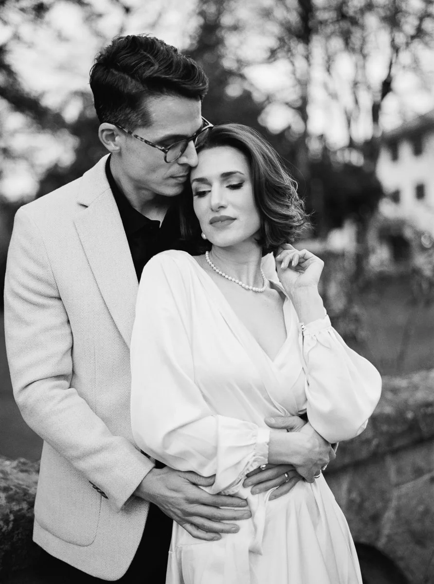 Black and white close-up of couple embracing, woman with soft waves and pearl necklace, man in blazer behind her