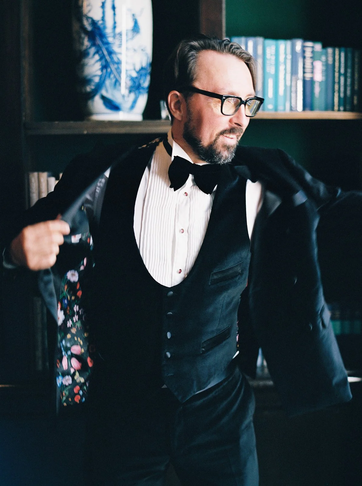Groom putting on tuxedo jacket in a library setting with bookshelves behind him.