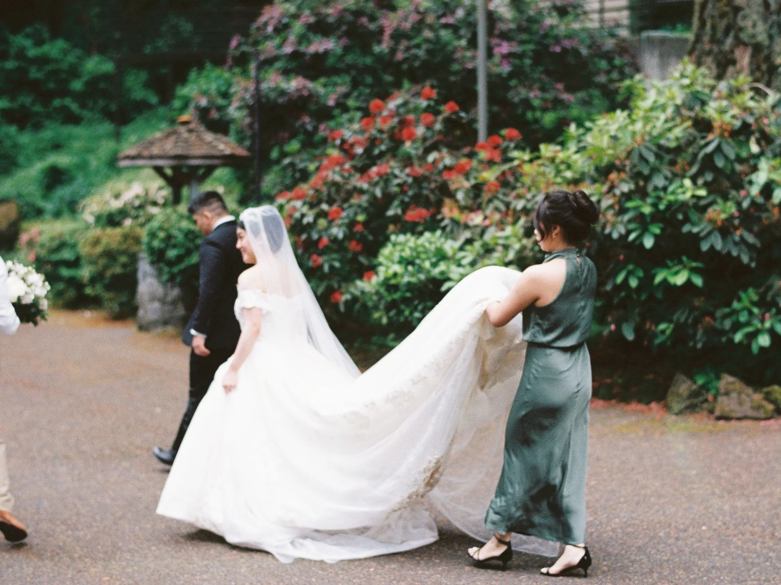 A bridesmaid in a forest green dress carries the bride's lace train through The Grotto gardens as the couple walks ahead, red rhododendrons blooming behind them