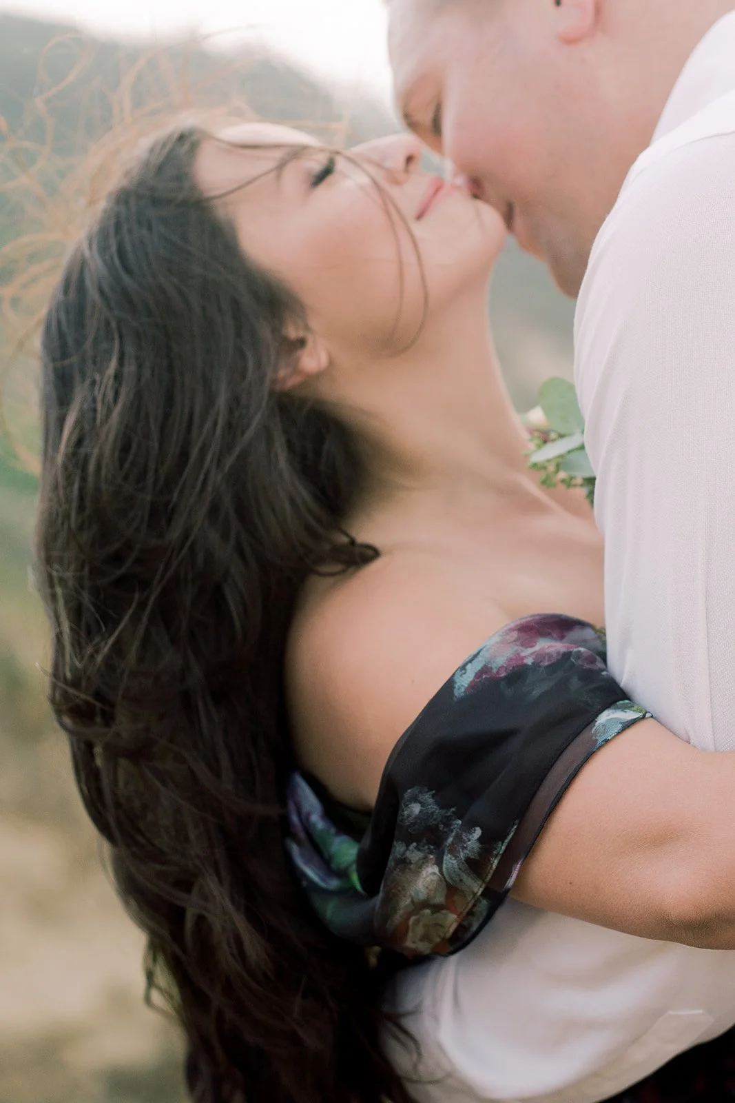 Close-up of couple kissing with flowing hair, muted coastal backdrop and soft, airy tones