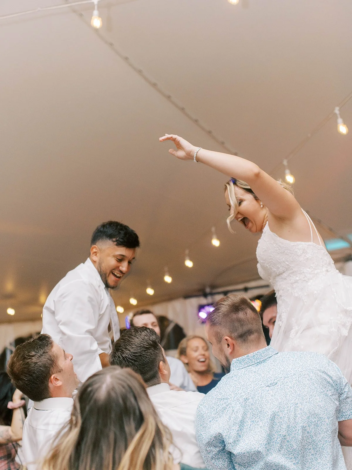 Bride and groom are raised on guests’ shoulders during the reception, celebrating beneath string lights in a wedding tent.