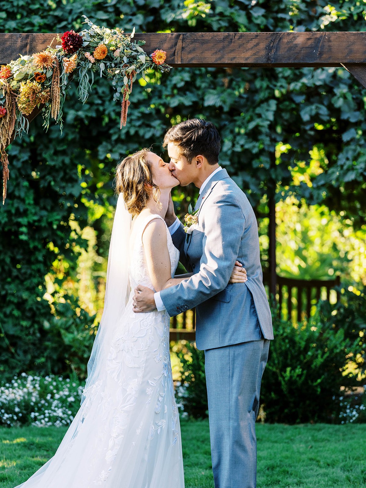 Bride and groom sharing first kiss under floral arch at outdoor garden wedding