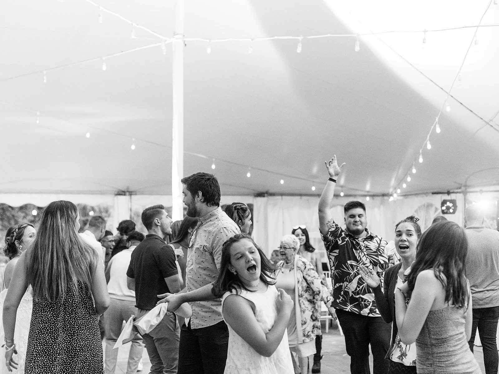 Wedding guests celebrate and dance together under a white reception tent in black-and-white.