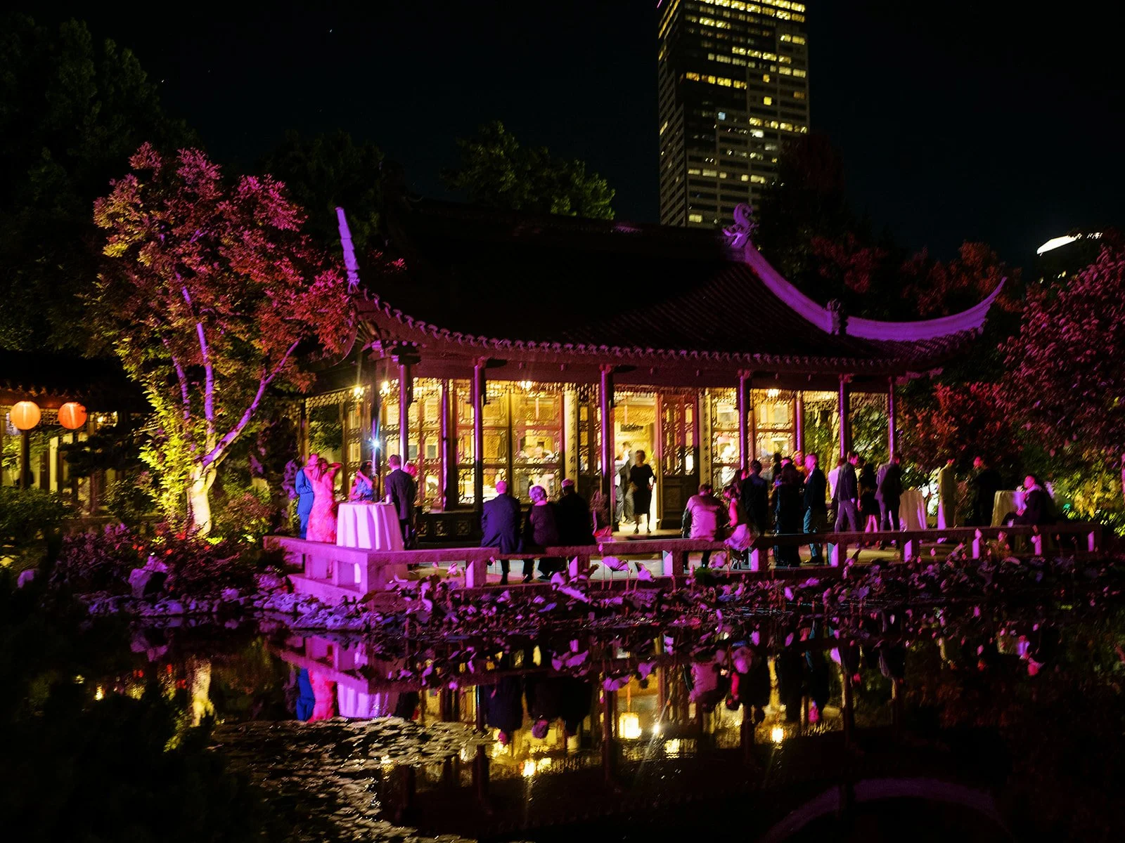 Guests gather on the illuminated lakeside terrace under purple and gold evening lighting, the pavilion's curved roofline and city high-rise reflected in the still water below a starry sky.
