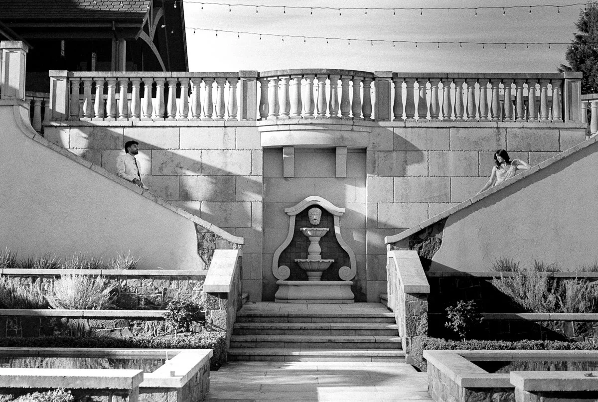 Black-and-white wide shot of South Asian couple posed across stone courtyard staircases with fountain backdrop.