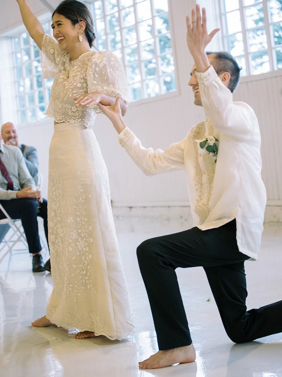 Groom kneeling barefoot on the dance floor with arm raised while the bride laughs and raises her arm during their energetic first dance, guests cheering in the background
