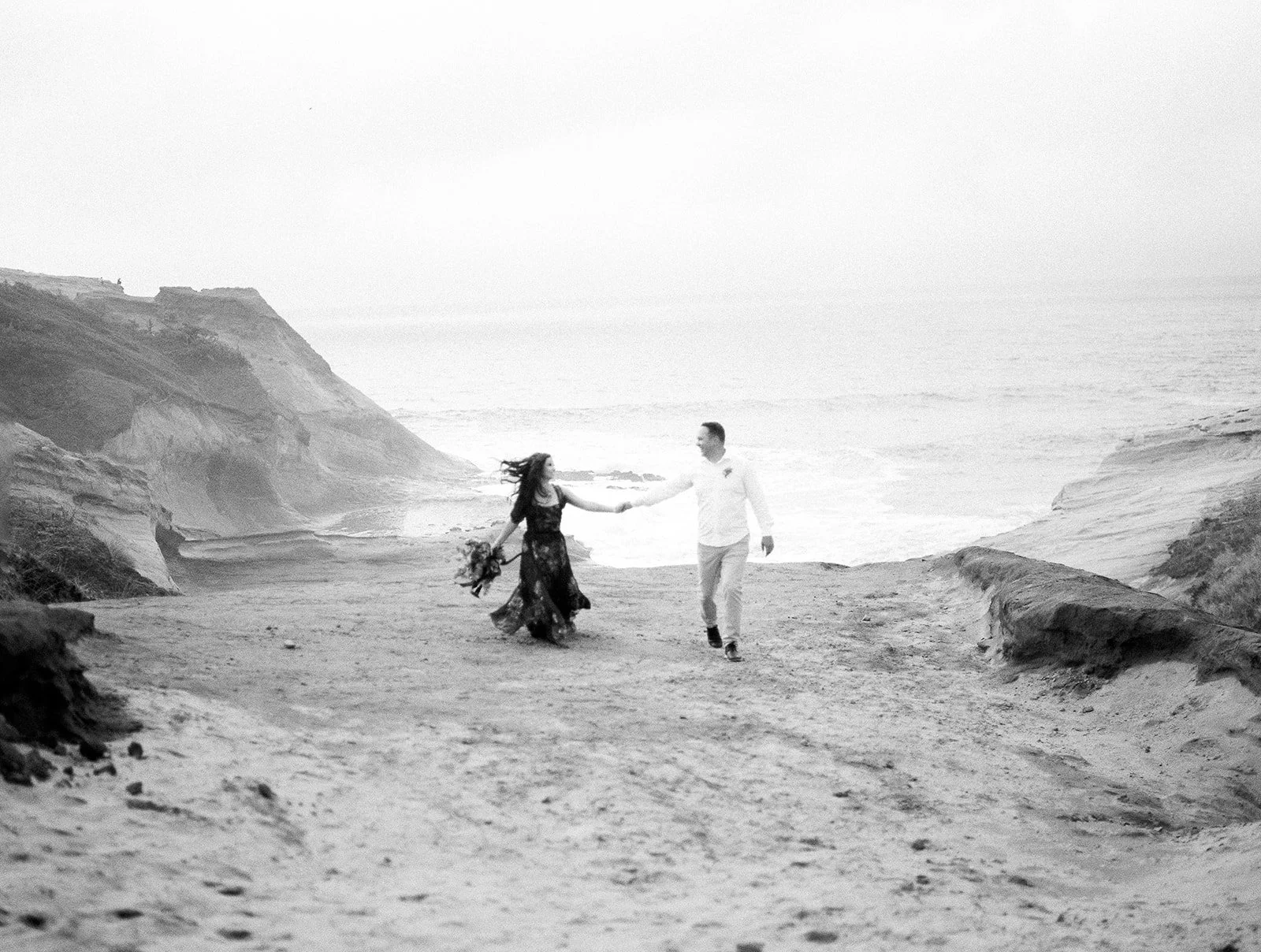 Couple holding hands and walking along coastal cliff overlooking ocean in soft, misty light