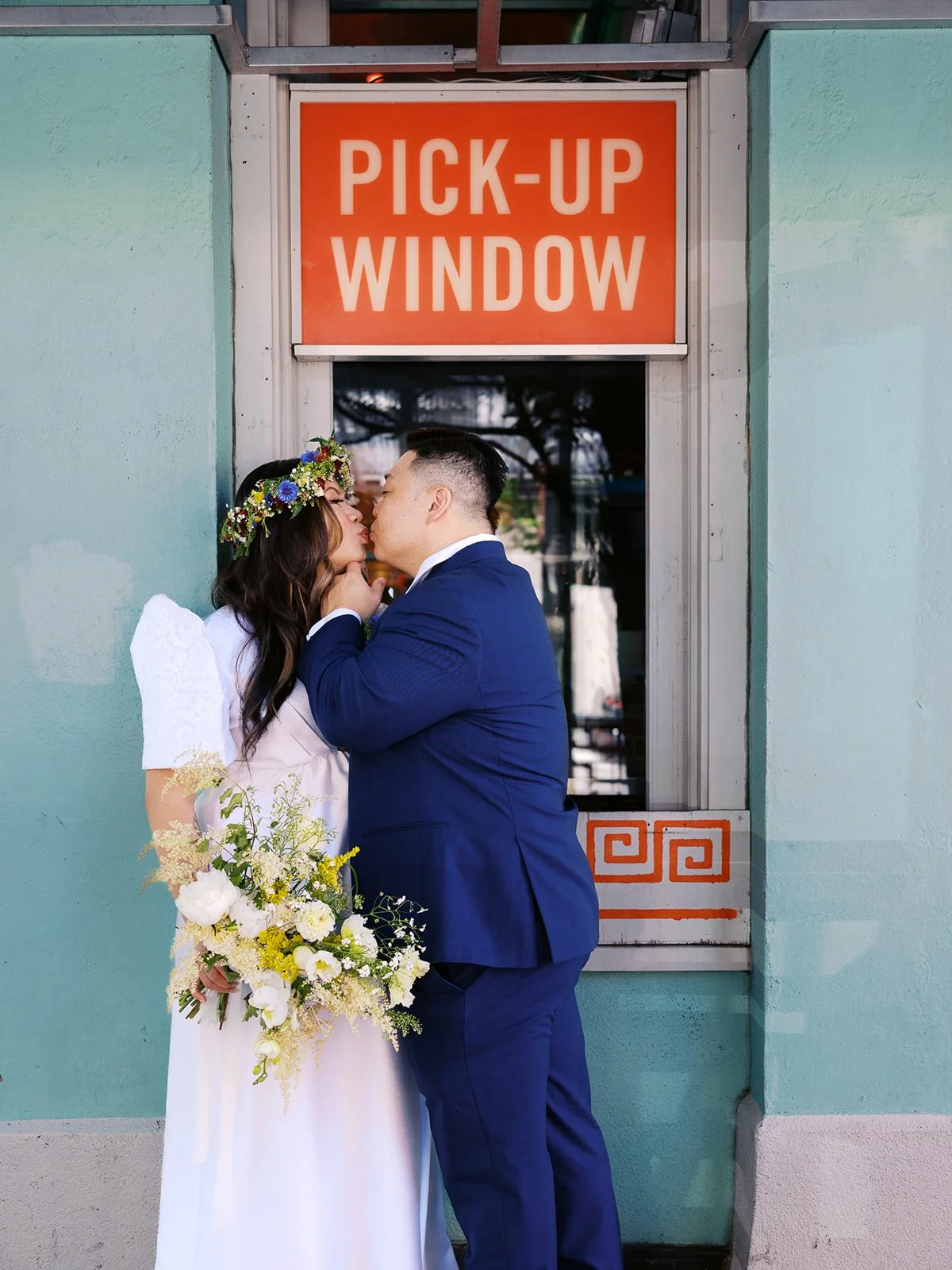 Newlyweds kiss beneath an orange pick-up window sign, with the bride holding her white and yellow wedding bouquet.