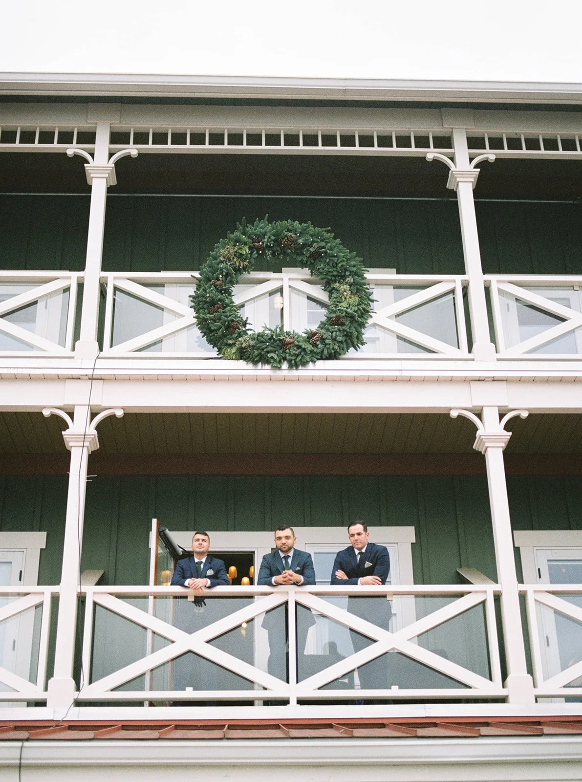 Groom and groomsmen lean along a white balcony beneath an oversized evergreen wreath.