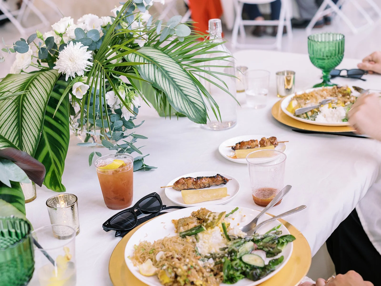 Reception table with Filipino-inspired food including chicken skewers, rice, and vegetables alongside a tropical floral centerpiece and the bride's cat-eye sunglasses
