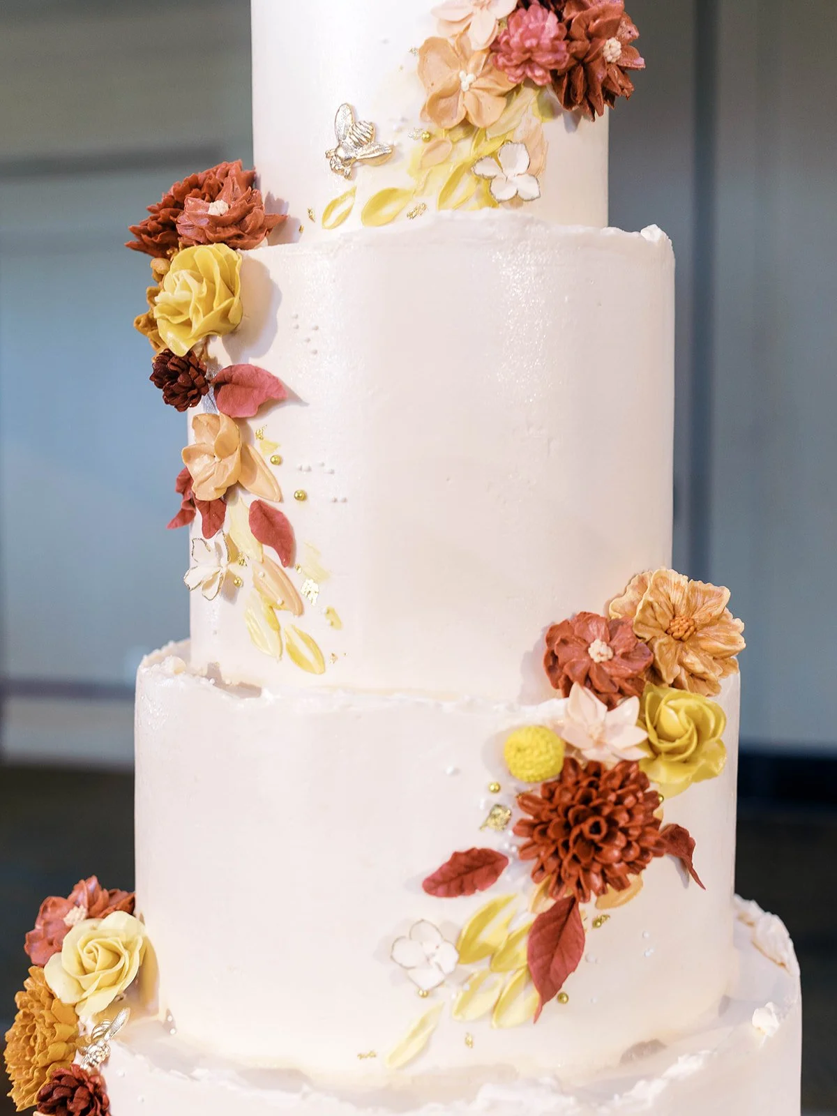 Four-tier white wedding cake decorated with sculpted flowers in rust, yellow, blush and cream tones.