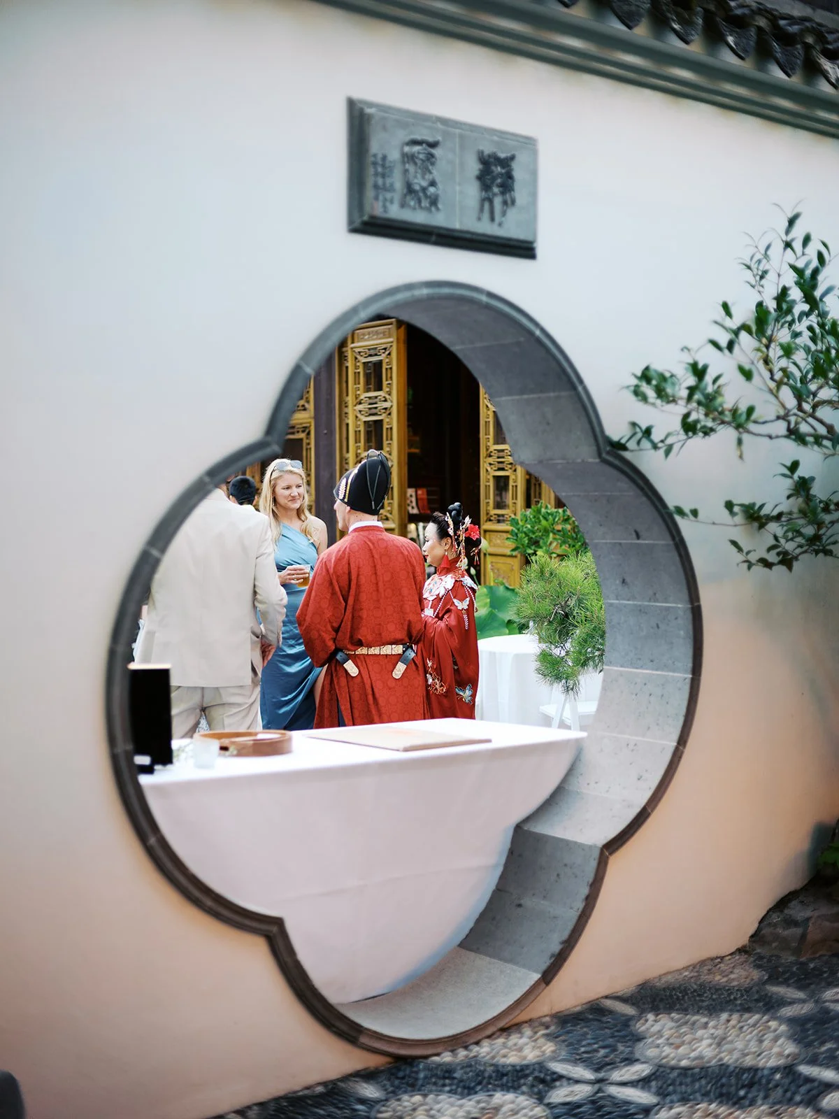The couple in red wedding robes framed within a circular cloud-collar moon gate, chatting with guests, the ornate golden hall interior visible behind them.