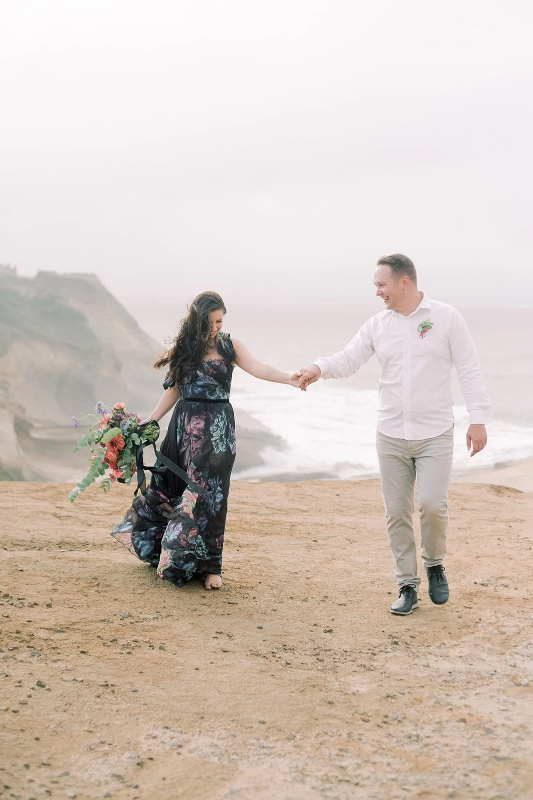 Couple holding hands and walking along Oregon Coast cliff, woman carrying bouquet, overcast sky