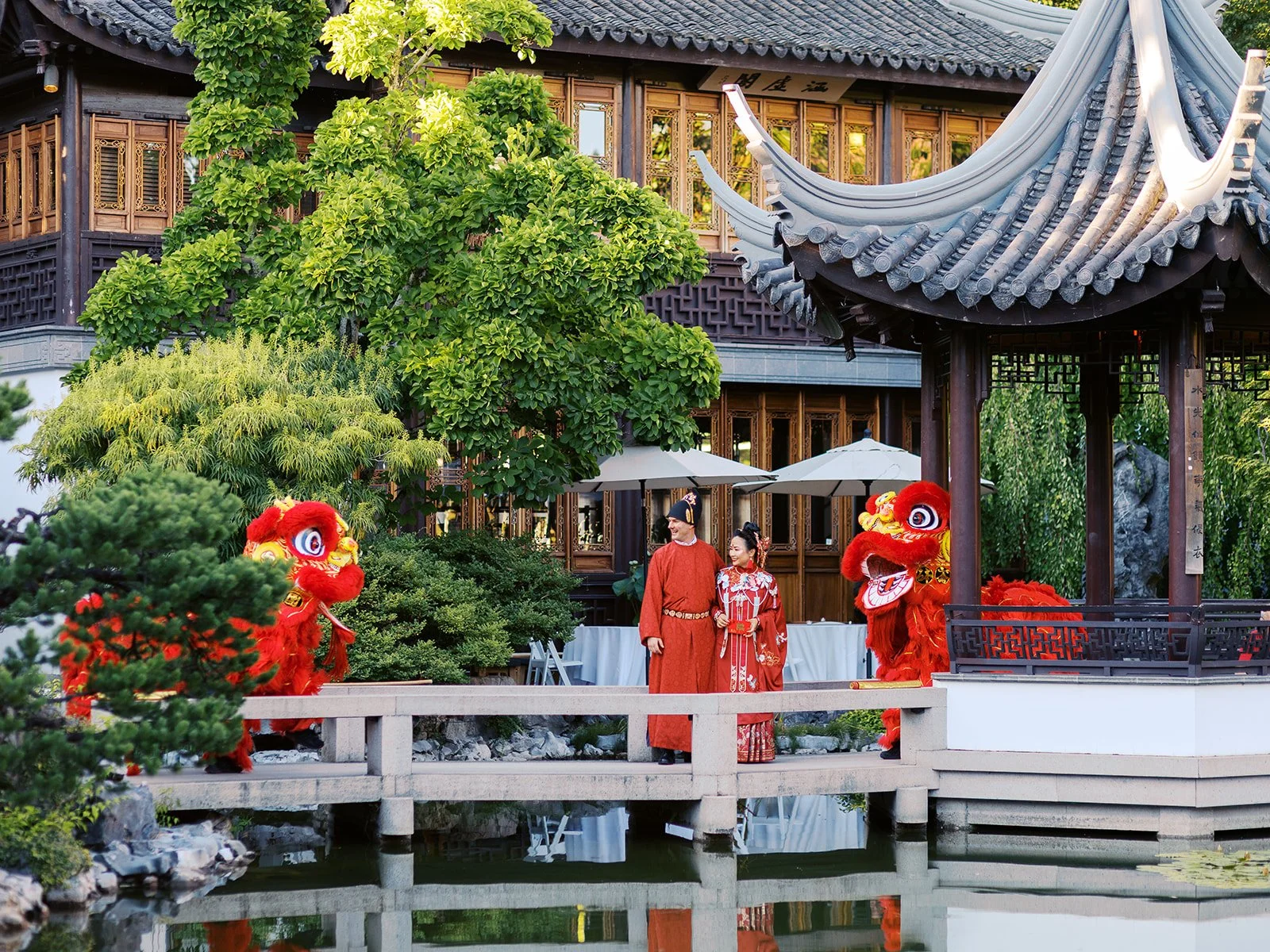 Wide shot of the couple in red wedding robes standing on the bridge flanked by two red lion dance costumes, the full garden and pavilion architecture reflected in the lake below.