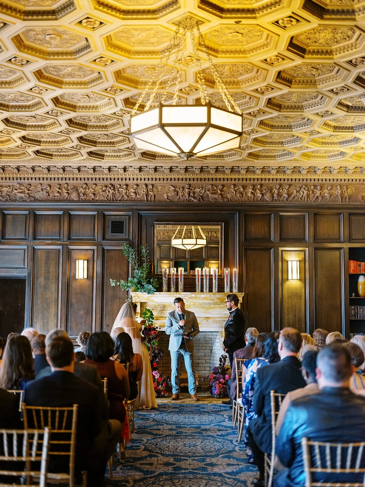 Wide view of wedding ceremony with guests seated under a patterned ceiling and chandeliers.