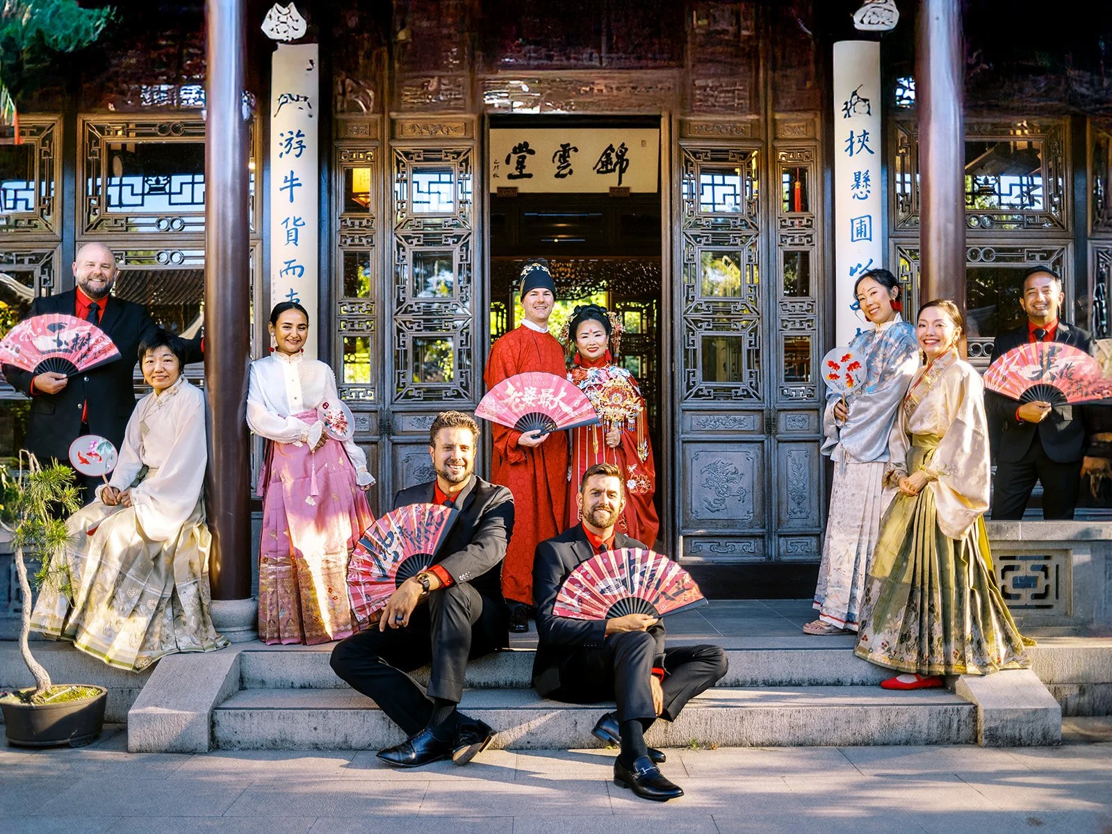 Full wedding party posing with red fans on the steps of an ornately carved wooden pavilion at Lan Su Chinese Garden Portland