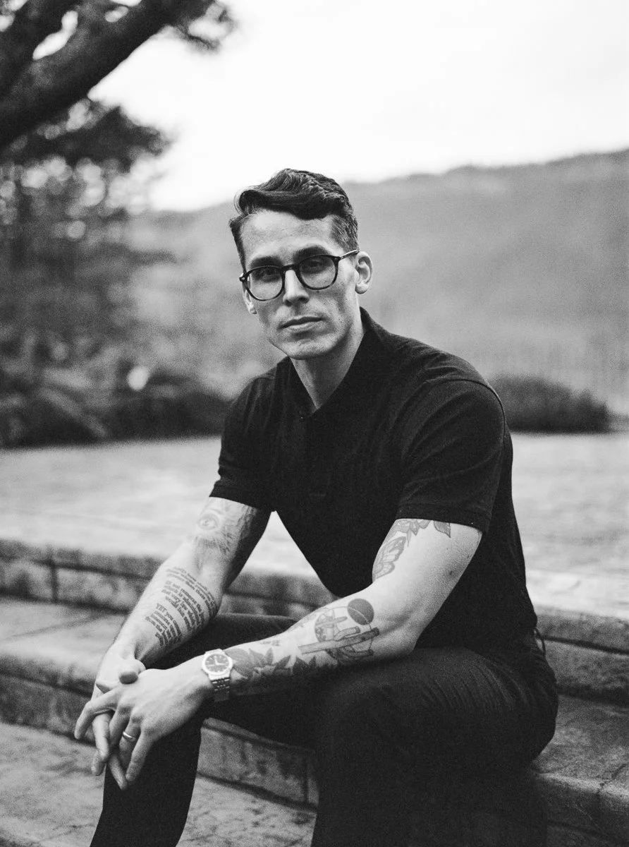 Black and white portrait of a tattooed man in glasses and a black polo seated on stone steps with a mountain backdrop.