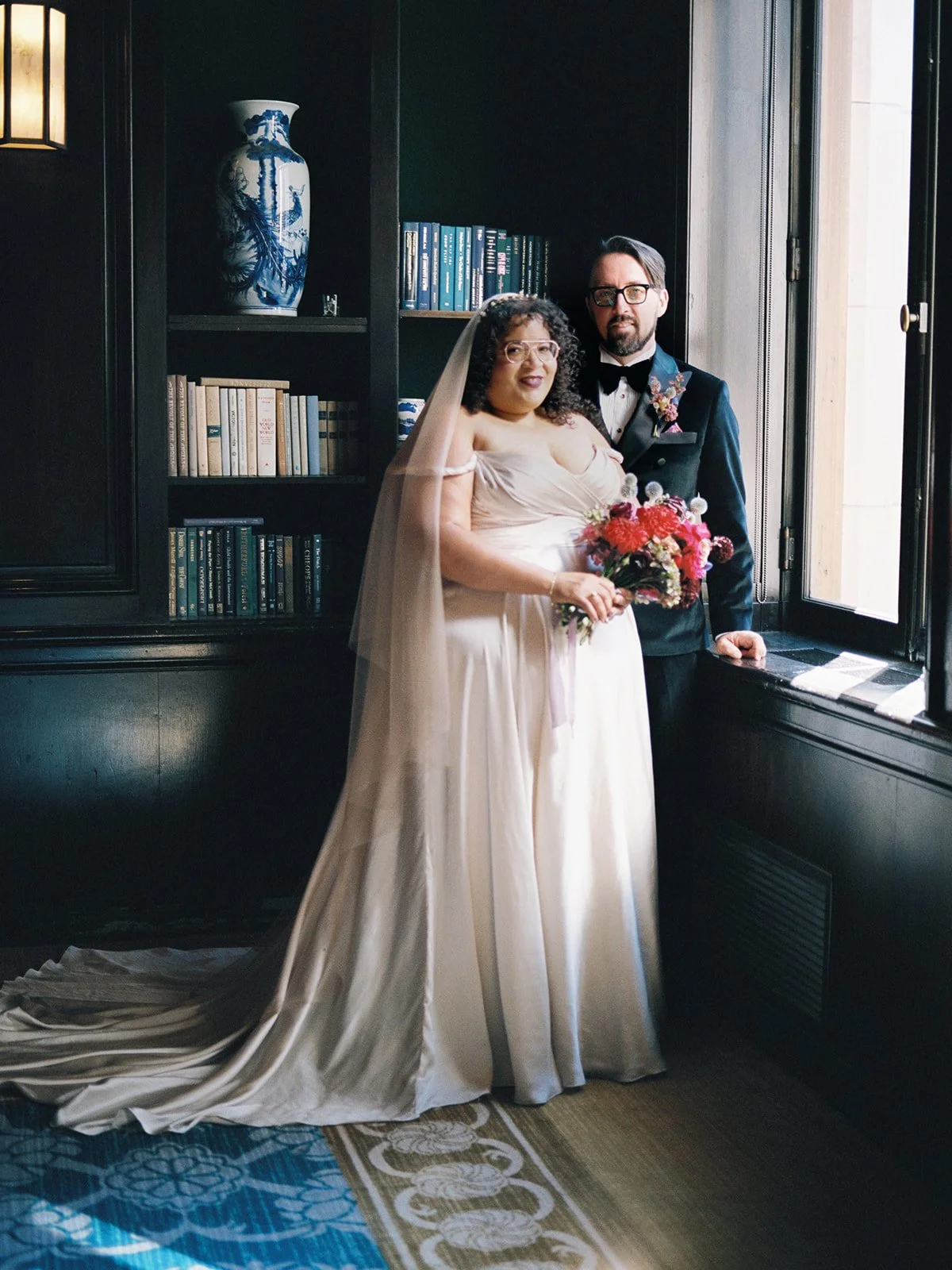 Bride and groom standing by window in library-style room, holding bouquet with shelves and blue ceramic vase behind.