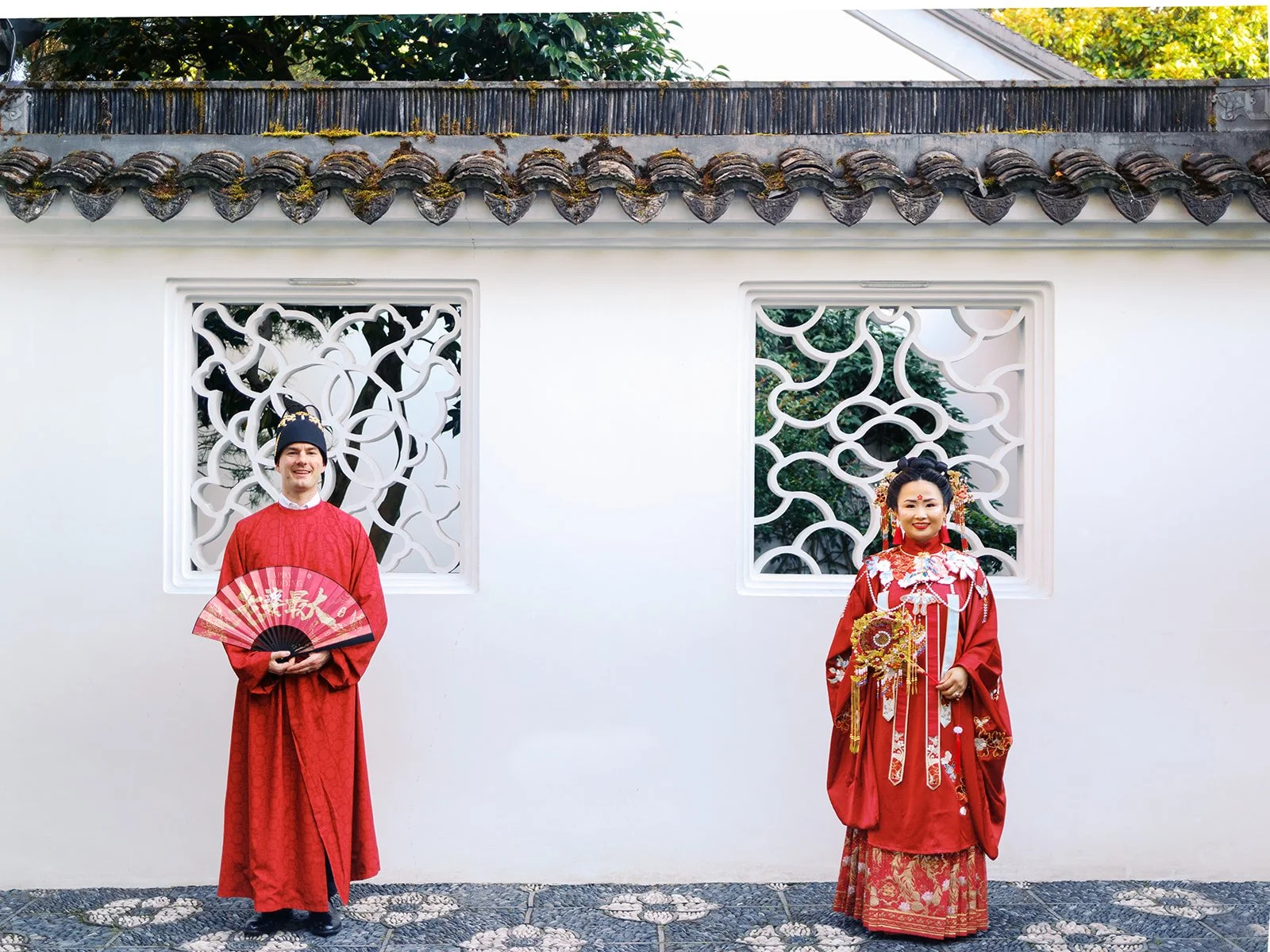 Bride and groom standing in separate decorative lattice window frames against a white garden wall, both in full red traditional Chinese wedding attire