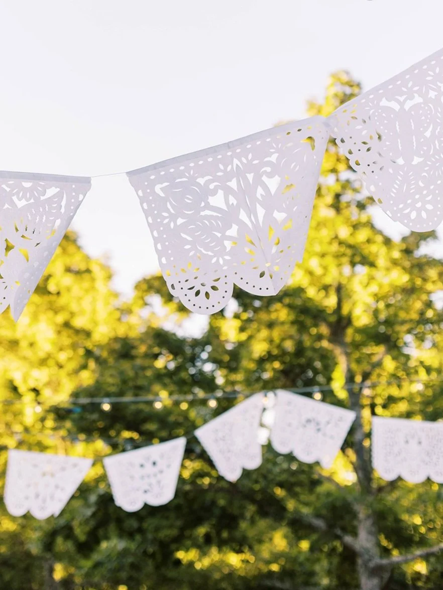 White papel picado banners hang outdoors among trees and string lights at a wedding celebration.