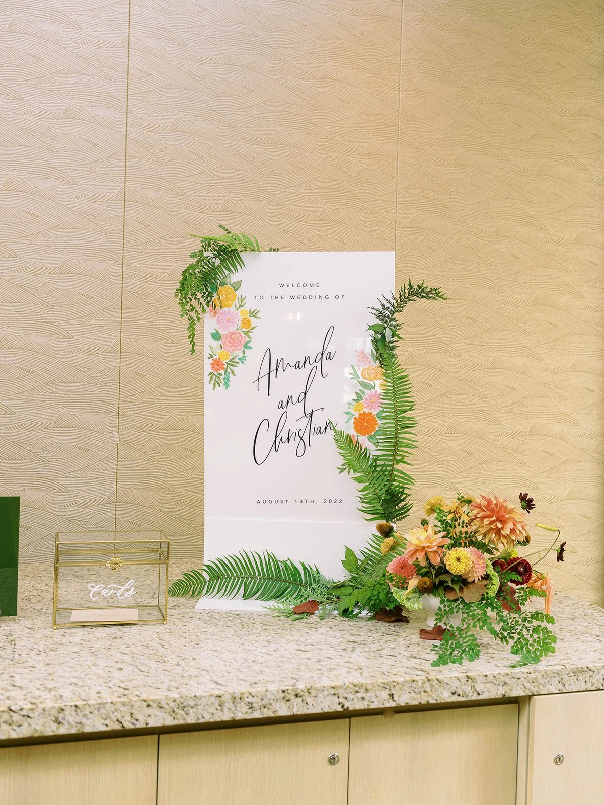 Wedding welcome sign decorated with illustrated florals and real fern fronds, displayed on a granite surface alongside a gold card box and a colorful flower arrangement