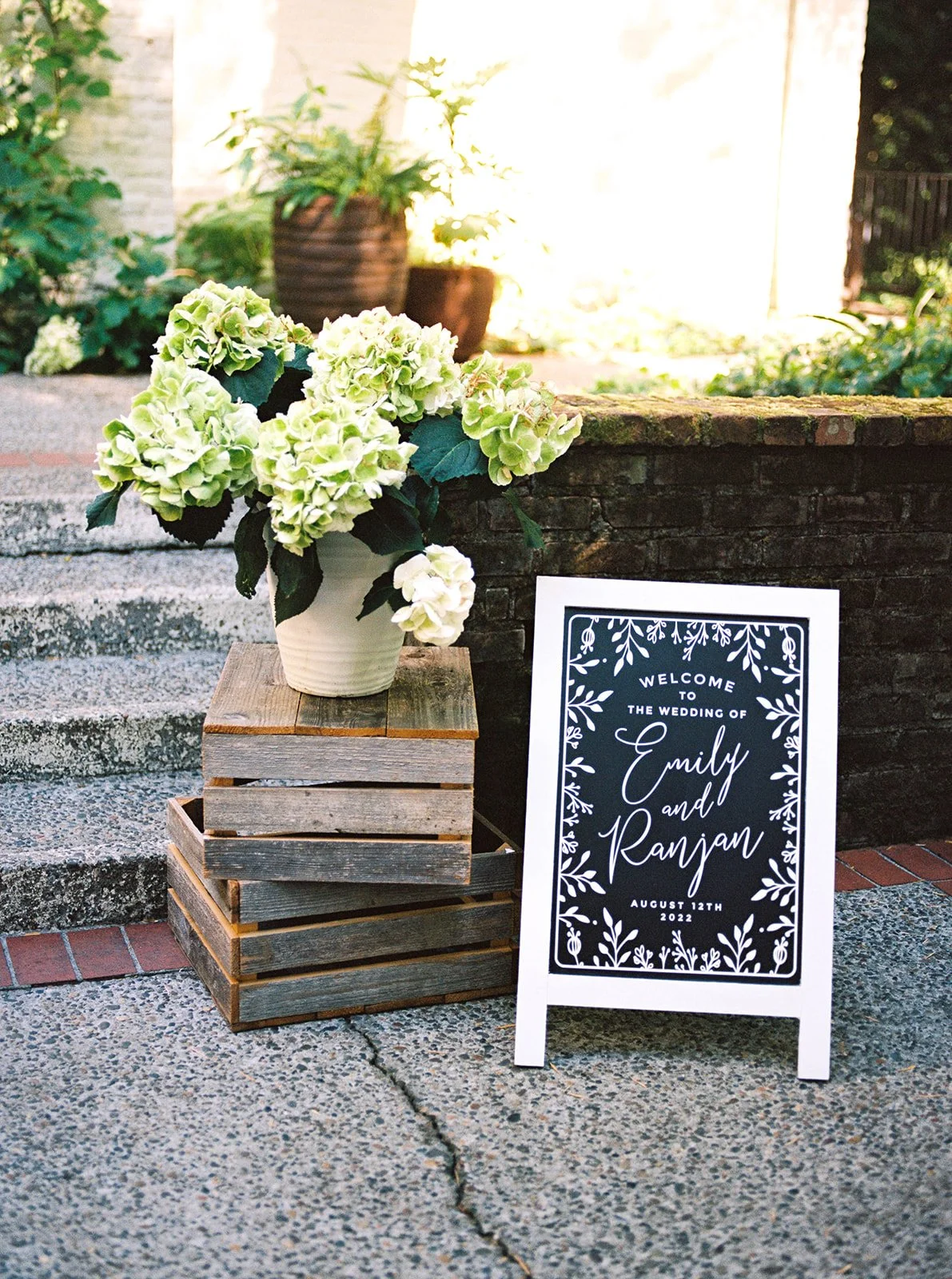 Welcome sign for Emily and Ranjan’s wedding displayed beside hydrangeas and wooden crates.