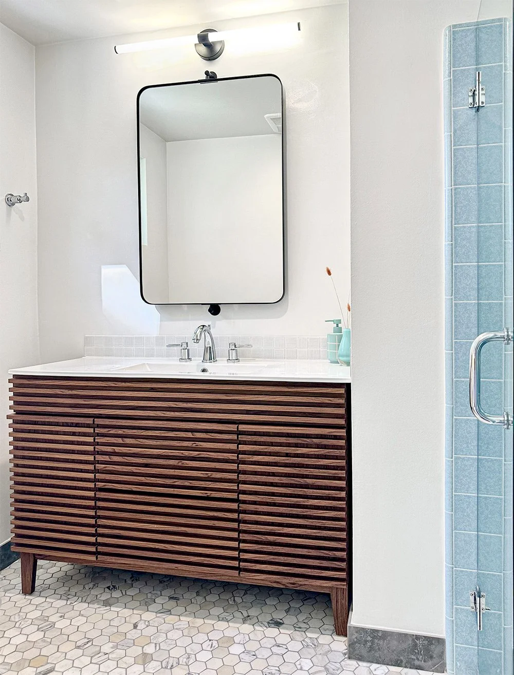 A modern bathroom vanity with a wooden slatted cabinet, white countertop, two sinks, and a rectangular wall mirror illuminated by a horizontal light fixture. A small vase with dried plants and a soap dispenser are on the countertop. To the right, there is a shower with blue tiles and a glass door.