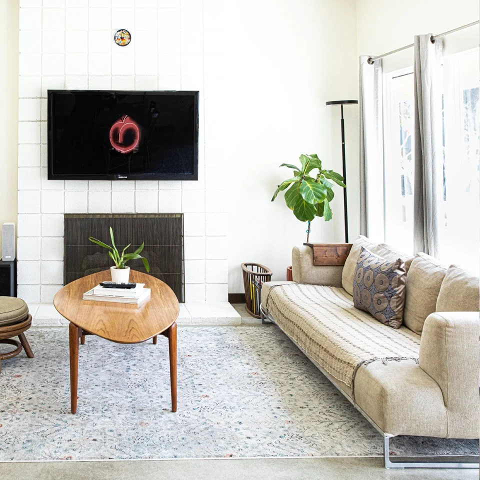 Living room with a wall-mounted flat-screen TV, a wooden coffee table with a potted plant, a beige sofa with pillows, large windows with curtains, a tall floor lamp, and a patterned area rug.