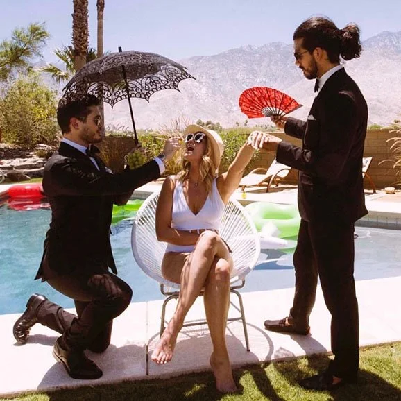 A woman in a white swimsuit sitting by a pool with two men in black suits, one kneeling and the other standing, around her in a sunny outdoor setting with mountains in the background.
