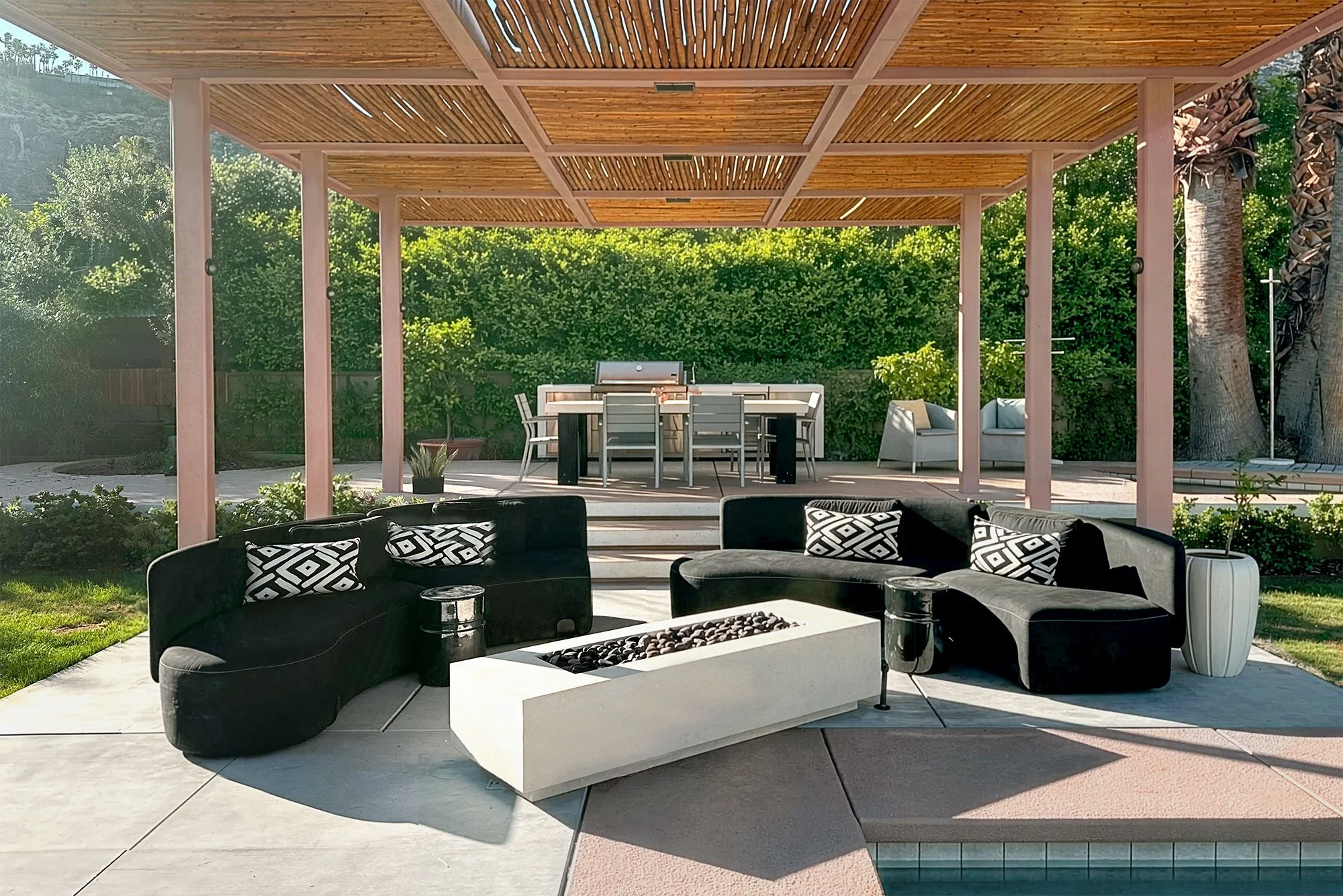 Outdoor patio with black sofas, decorative pillows, and a white rectangular fire feature in the foreground. In the background, a wooden pergola with seating and a lush green hedge.
