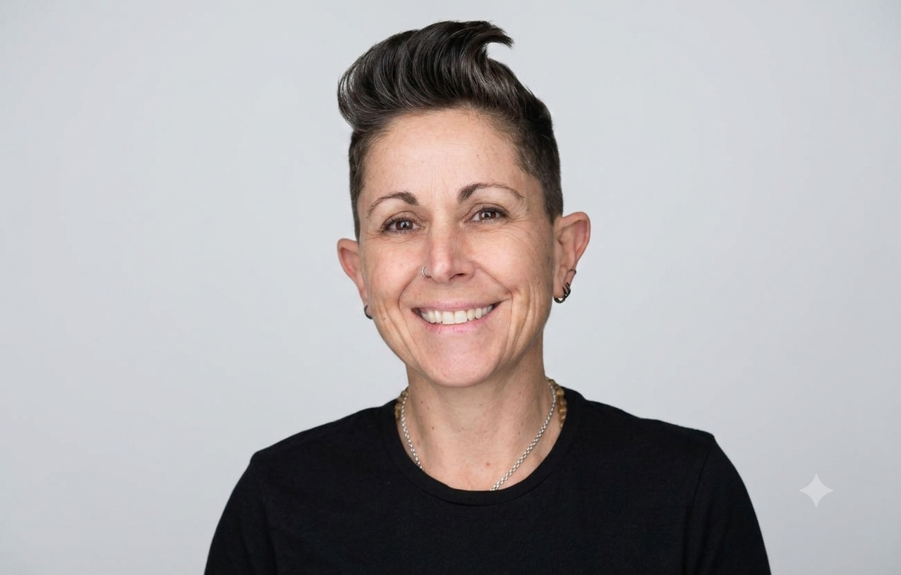 Smiling woman with short, styled hair wearing a black top, small hoop earrings, a nose ring, and a necklace, standing against an orange background.