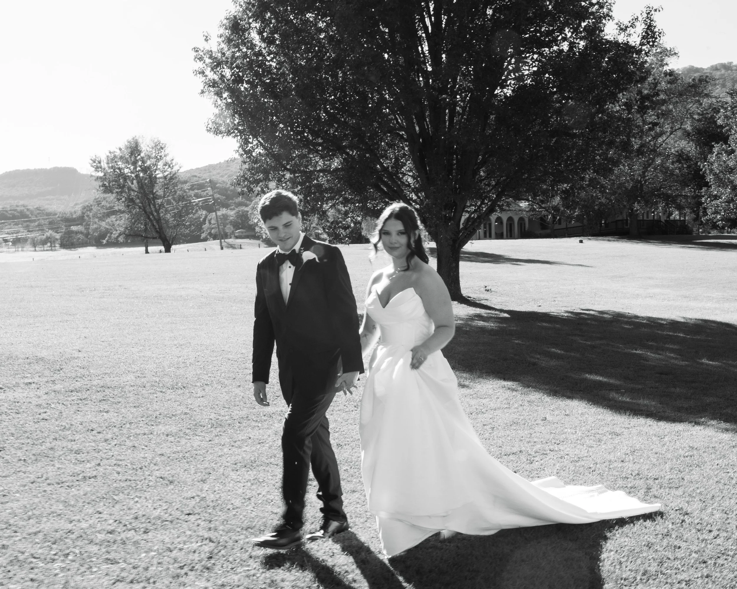 A black-and-white photo of a bride and groom walking outdoors on a sunny day. The bride is wearing a strapless white wedding gown, and the groom is dressed in a black tuxedo with a bow tie. They are holding hands and walking across a grassy area with a large tree in the background.