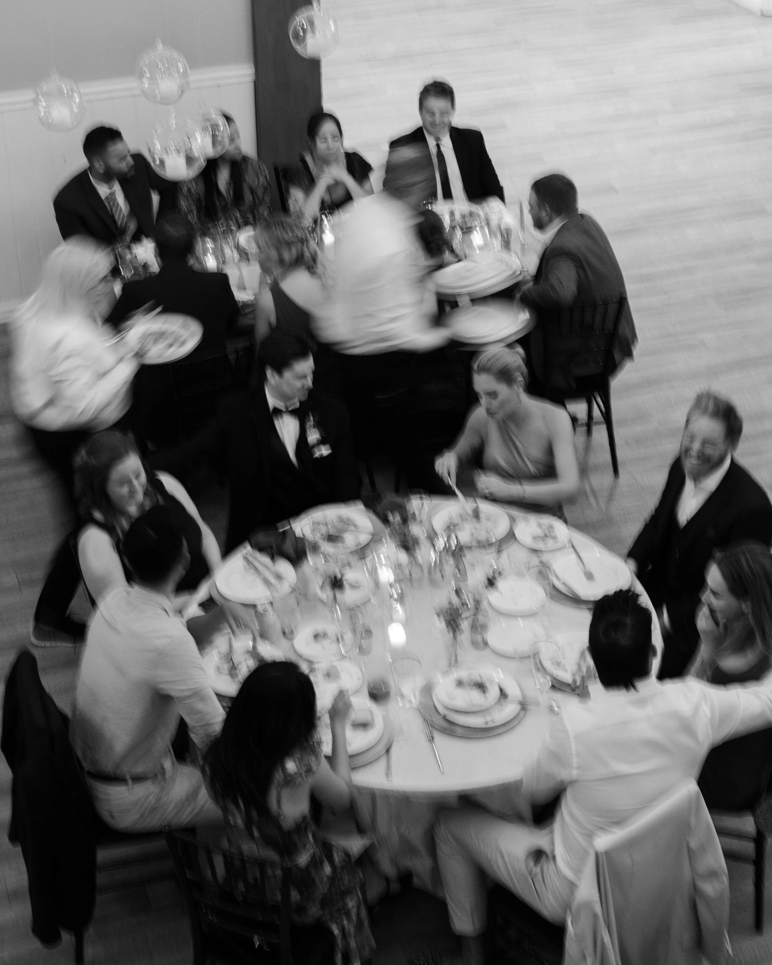 Black-and-white photo of people at a formal dinner party, sitting around multiple tables, with some guests eating and others mingling, in a well-lit room with balloons hanging from the ceiling.
