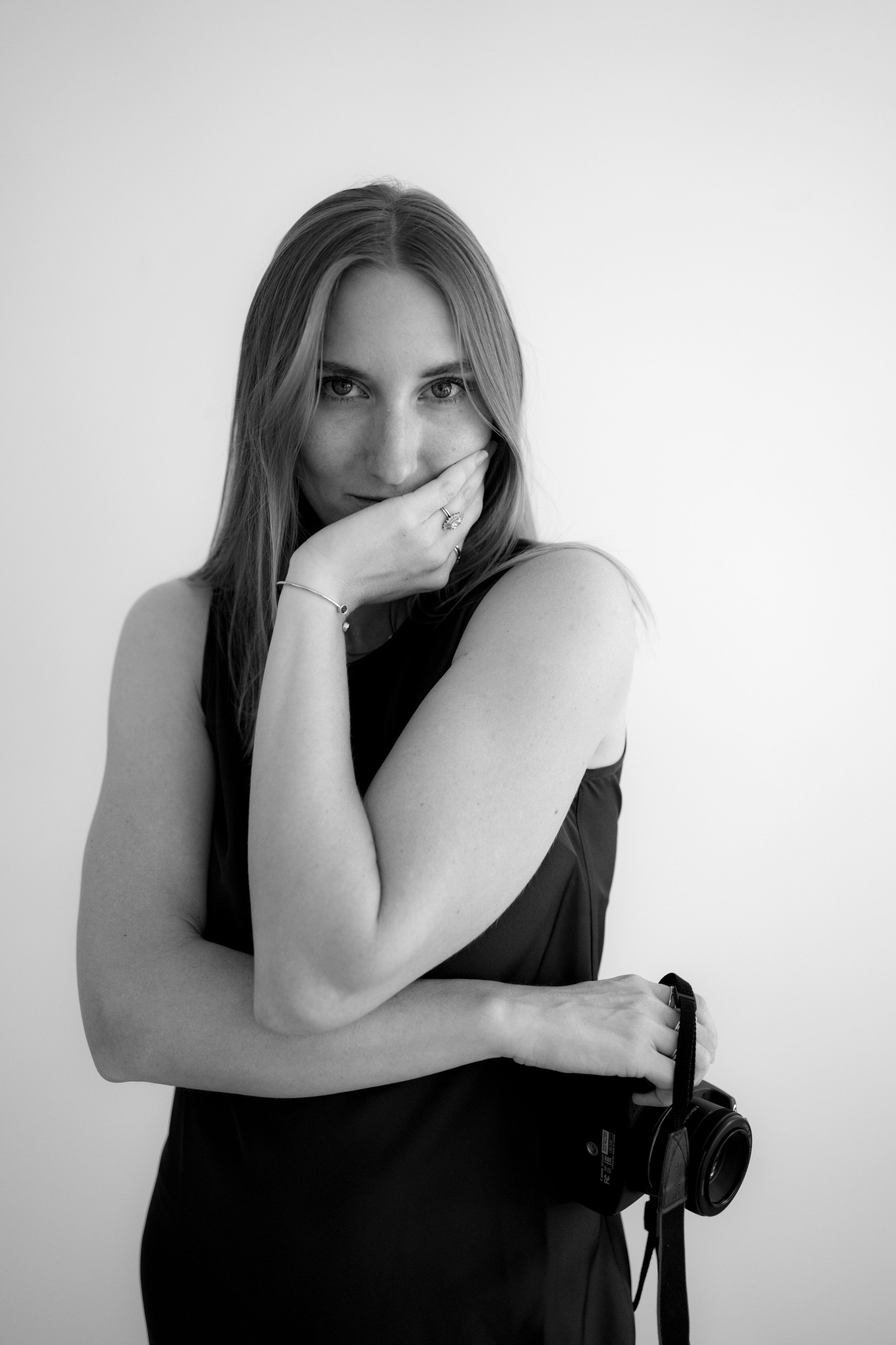 A woman with long hair holding a camera, partly covering her face with her hand, looking directly at the camera, in black and white.